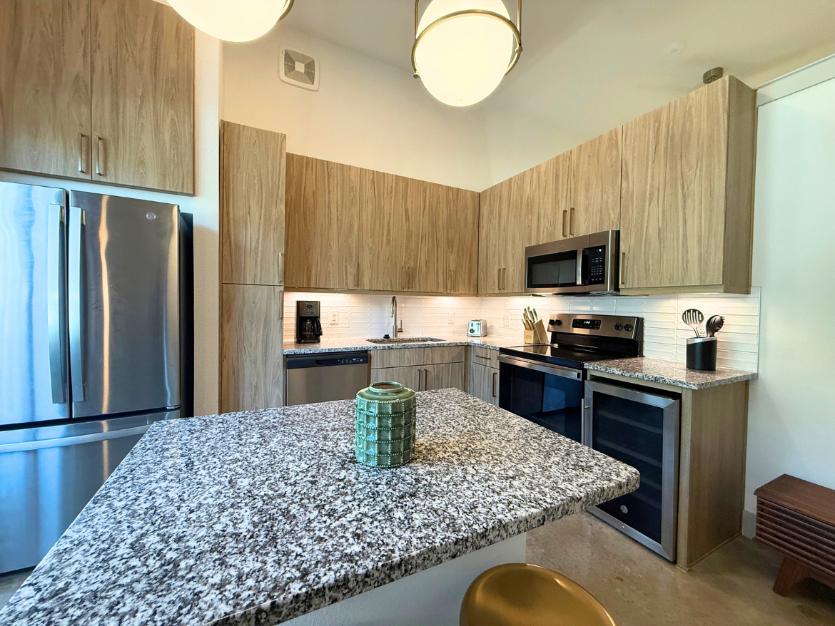 A modern kitchen featuring light wood cabinetry and a spacious granite countertop. The kitchen includes stainless steel appliances, such as a refrigerator, oven, and microwave. A small coffee maker is positioned near the sink, and utensils are neatly arranged in a holder. Under-cabinet lighting illuminates the backsplash, enhancing the overall brightness of the space. A decorative green vase sits on the countertop, adding a touch of color. The flooring is a polished concrete, and a wooden accent piece is visible in the corner.