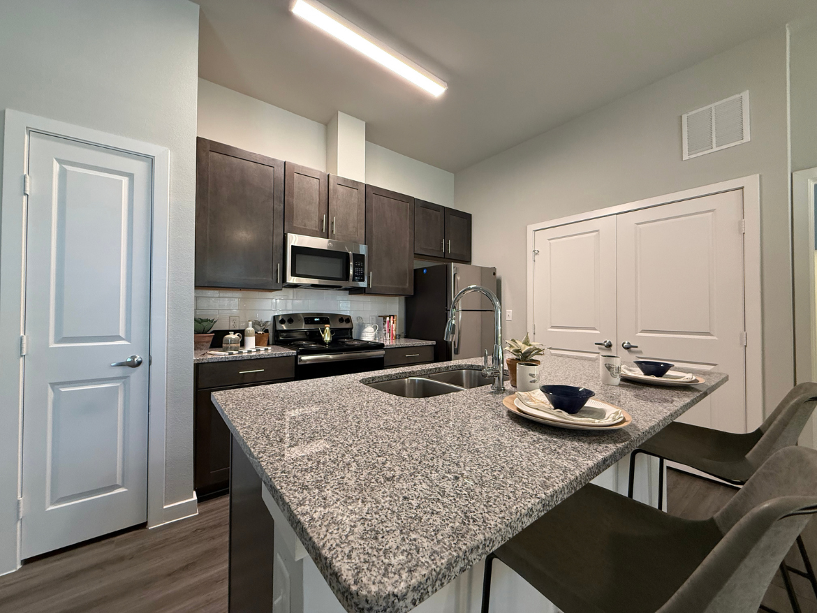 A modern kitchen features dark wood cabinetry and stainless steel appliances, including a microwave and stove. The countertop is made of gray granite, with a double sink and a sleek faucet. Two bar stools are positioned at the counter, which is set with plates and bowls. A small potted plant adds a touch of greenery. A door leads to another room, and there is a second set of double doors visible on the right side of the image. The flooring is a light wood laminate, contributing to the contemporary aesthetic.