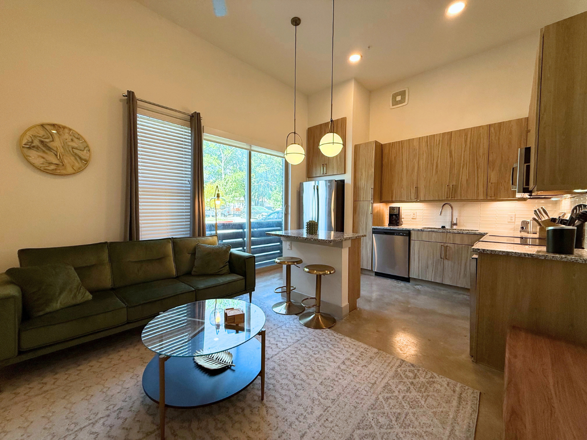 A modern living space featuring a green sectional sofa with cushions, positioned next to a round glass coffee table. The table has a decorative item on it, resembling a leaf. Behind the sofa, large windows allow natural light to flood the room, revealing a view of outdoor greenery. To the right, a kitchen area showcases wooden cabinetry, stainless steel appliances, and a granite countertop with bar stools. The flooring is polished concrete, and a light-colored area rug adds warmth to the space. Two pendant lights hang above the kitchen counter, enhancing the contemporary aesthetic.