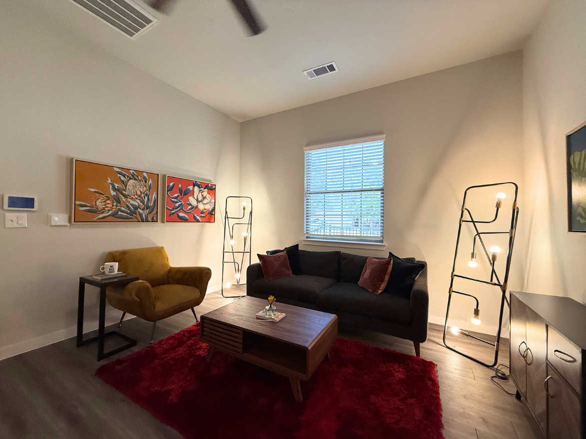 A cozy living room features a dark gray sectional sofa adorned with red and brown accent pillows. A wooden coffee table sits in front, with a small flower vase on it. To the left, a mustard-colored armchair complements the space, accompanied by a small black side table holding a mug. The wall displays two framed artworks with floral designs. A tall, modern floor lamp with multiple bulbs stands in the corner, providing additional lighting. Natural light filters through a window with blinds, and a plush red rug adds warmth to the wooden floor.