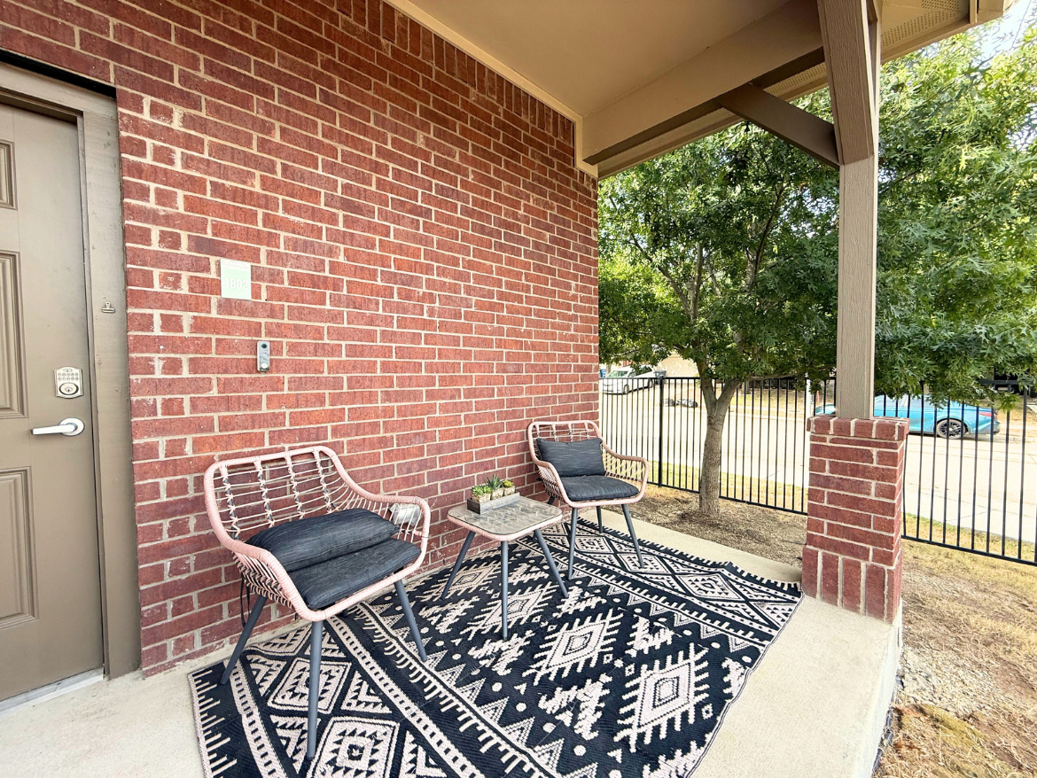 A cozy outdoor seating area features two stylish chairs with dark cushions, positioned on a patterned black and white rug. The backdrop is a brick wall with a door and a small decorative planter on a table between the chairs. Nearby, a tree provides some greenery, and a fence encloses the area, with a glimpse of a car parked outside. The setting is inviting and well-maintained.