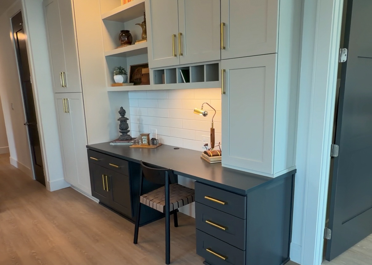 A modern workspace features a sleek, dark gray desk with three drawers and a matching chair with a patterned seat. Above the desk, there are light gray cabinets with gold handles, showcasing decorative items and a small plant. A white tiled backsplash is illuminated by a stylish desk lamp, and a small decorative tray holds various items, including a statue and books. The flooring is light wood, enhancing the contemporary aesthetic of the space.