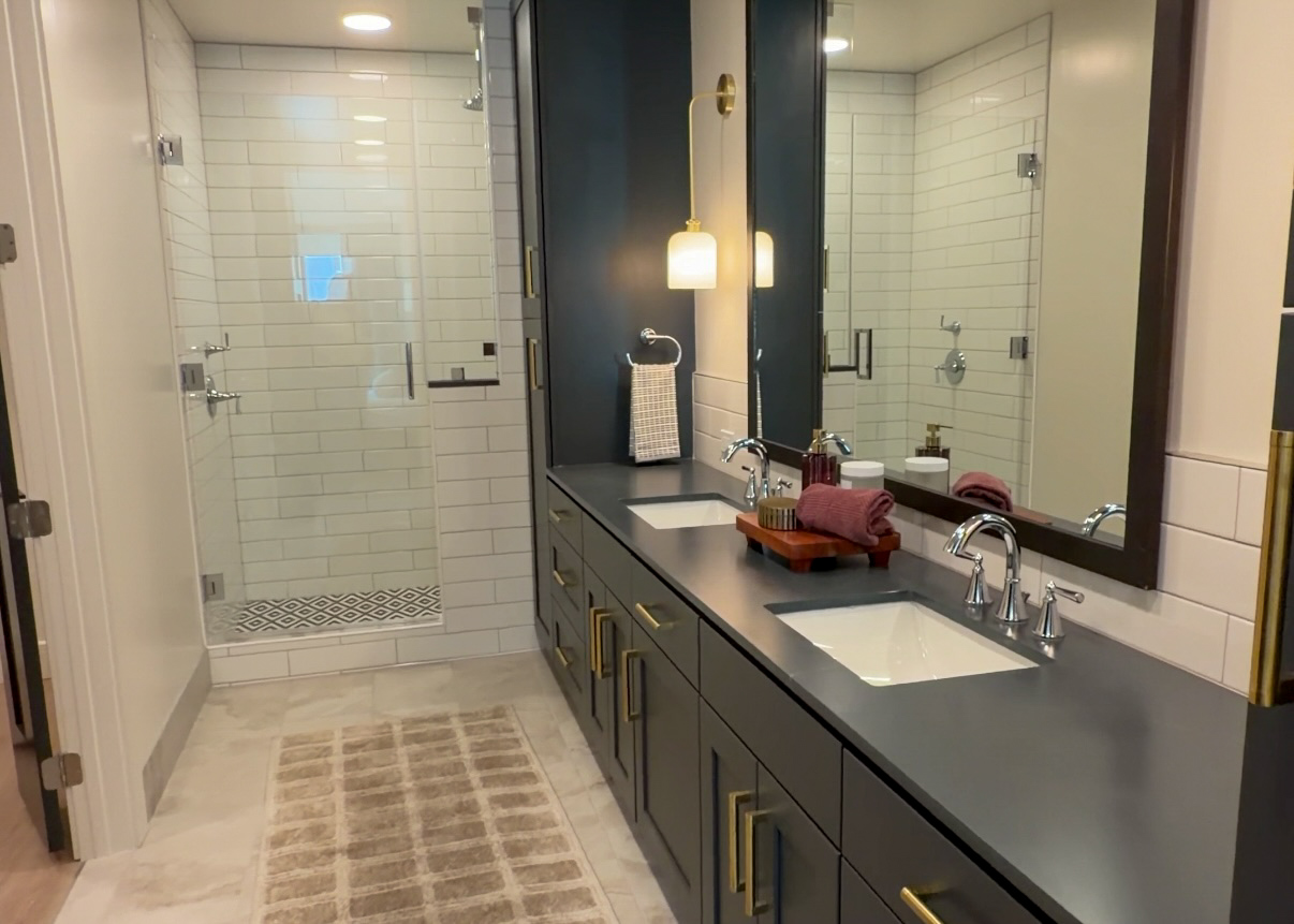 A modern bathroom features a spacious layout with a walk-in shower enclosed by clear glass and white subway tiles. The shower area has a decorative tile border at the bottom. A double vanity with a dark countertop holds two sinks, each with sleek chrome faucets. Above the vanity, there are large mirrors and a stylish wall-mounted light fixture. The vanity is accented with gold hardware and includes decorative items like rolled towels and a wooden tray. A soft, beige rug with a grid pattern lies on the floor, complementing the light-colored tiles.
