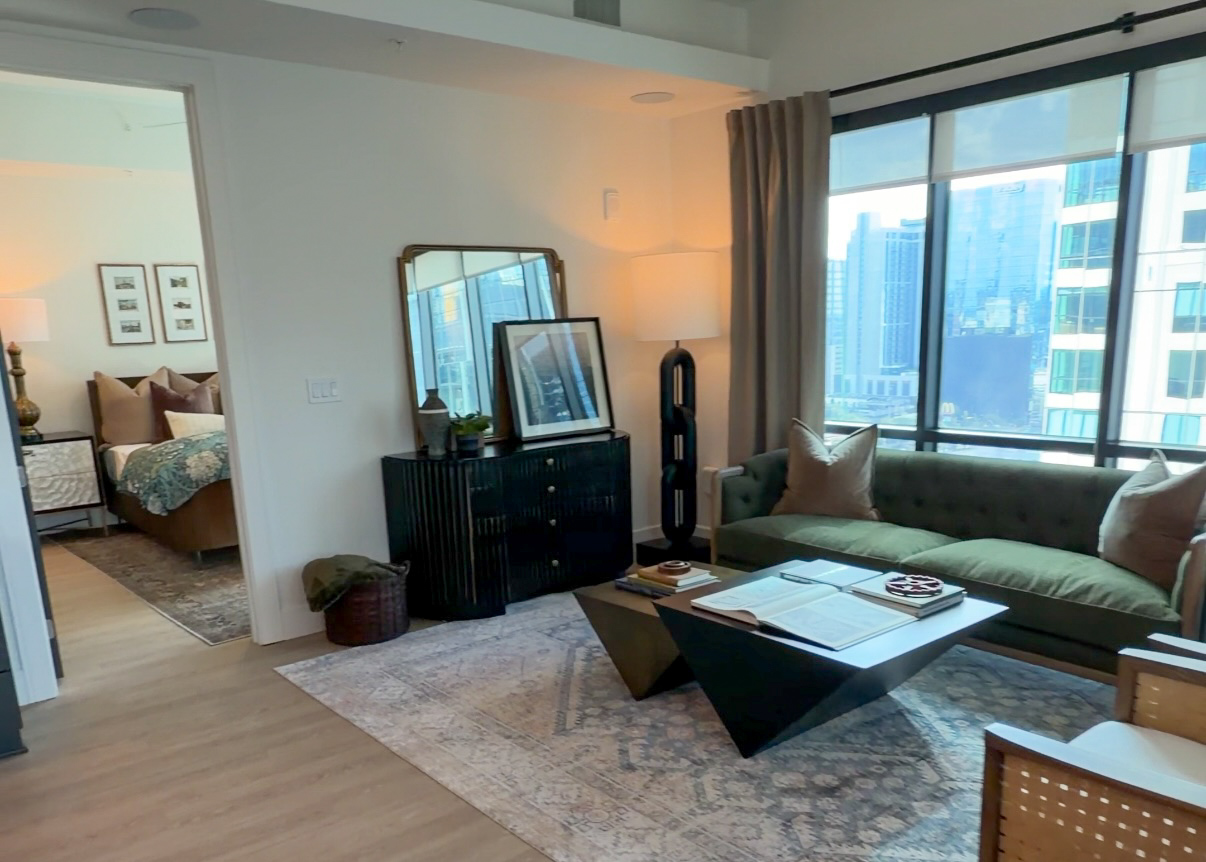 A modern living space features a green tufted sofa with decorative pillows, positioned next to a sleek black coffee table with a geometric design. On the table, there are books and a decorative item. A large area rug with intricate patterns lies beneath. To the side, a dark wooden cabinet holds a lamp and framed artwork, while a tall, slender lamp stands nearby. Large windows provide a view of the city skyline, and a doorway leads to a bedroom area with a bed adorned in patterned bedding.