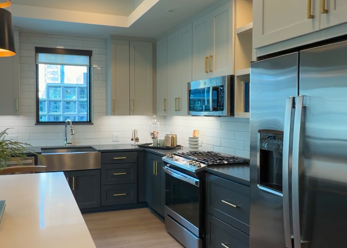 A modern kitchen featuring a combination of light and dark cabinetry. The upper cabinets are a soft gray with gold handles, while the lower cabinets are a darker shade. A stainless steel refrigerator and a gas stove with an oven are present, along with a built-in microwave above the stove. A large farmhouse sink is positioned beneath a window, allowing natural light to enter. The countertop is a sleek dark surface, and there are decorative items, including a small plant and dishware, on display. The backsplash consists of white subway tiles, enhancing the contemporary aesthetic.
