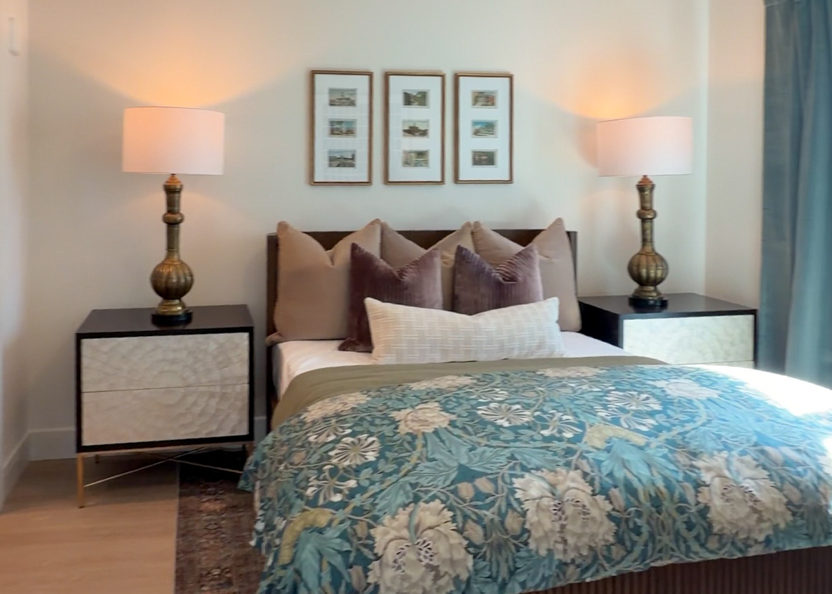 A cozy bedroom features a bed adorned with a colorful floral quilt in shades of blue and cream. The bed is flanked by two stylish nightstands with textured fronts and gold accents. Each nightstand holds a decorative lamp with a round white shade and a metallic base. Behind the bed, there are three framed pictures arranged in a row. The room is softly illuminated, creating a warm and inviting atmosphere, complemented by light blue curtains.