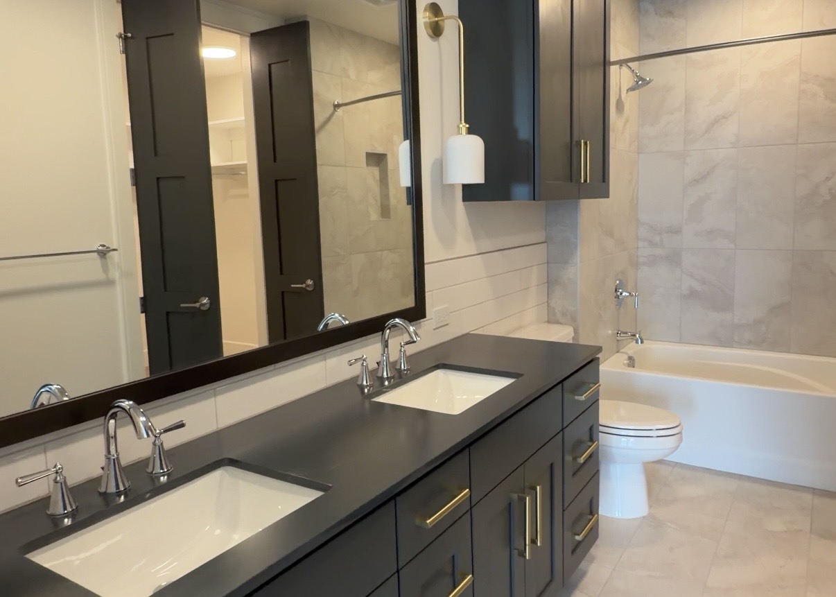 A modern bathroom features a double vanity with a dark countertop and two white sinks. Above the vanity, a large mirror reflects the space, which includes stylish wall-mounted lighting. The cabinetry is dark with gold hardware. To the right, a white bathtub is positioned against a wall with light-colored tiles, while a toilet is nearby. The overall design is sleek and contemporary, with a neutral color palette and clean lines.