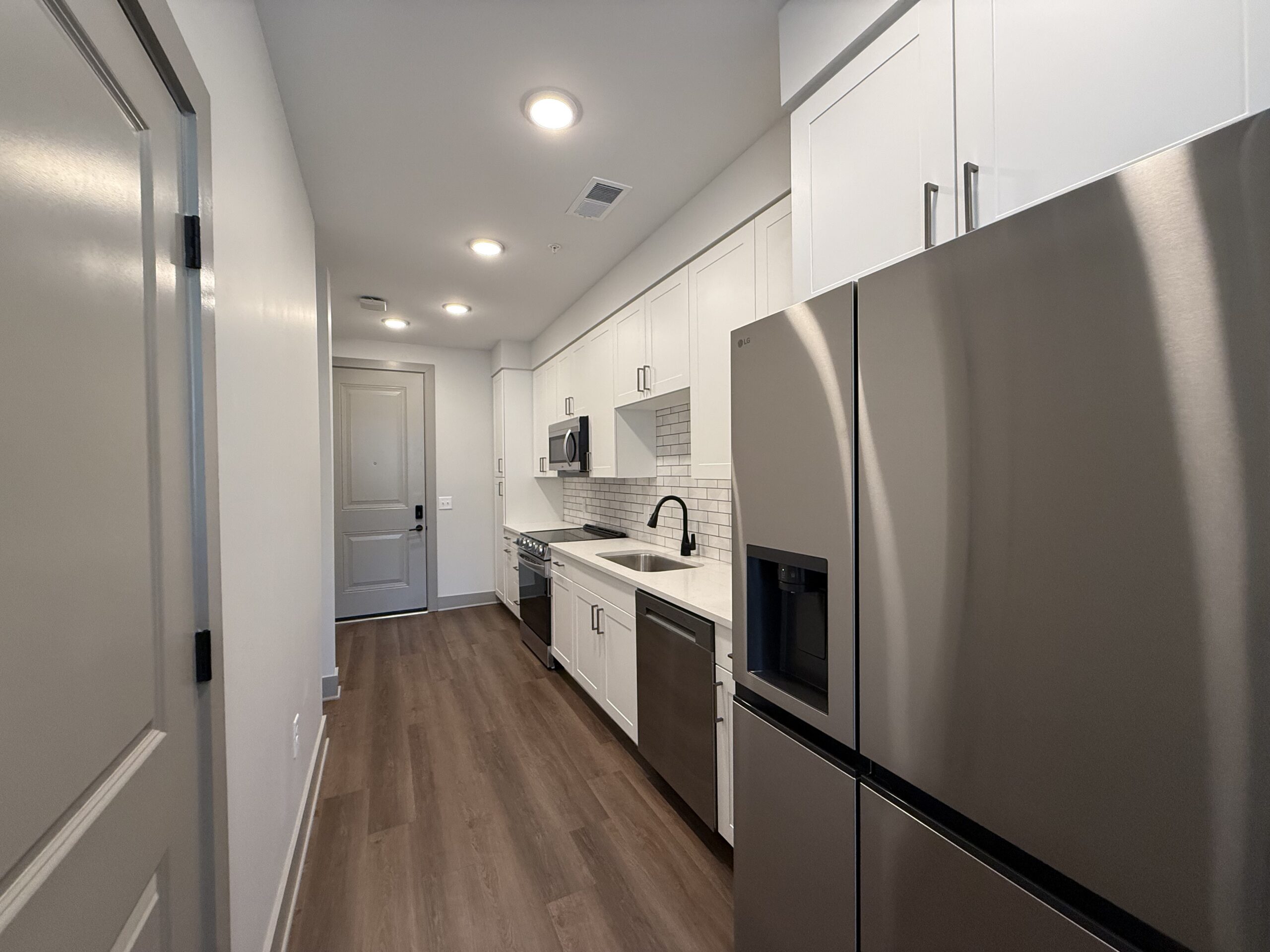 A modern kitchen features white cabinetry, stainless steel appliances, and a light-colored countertop. The layout includes a refrigerator, a microwave, a stove, and a sink with a black faucet. The backsplash consists of white subway tiles. The flooring is a warm wood tone, and there is a door leading to the outside at the end of the hallway. The space is well-lit with recessed lighting.