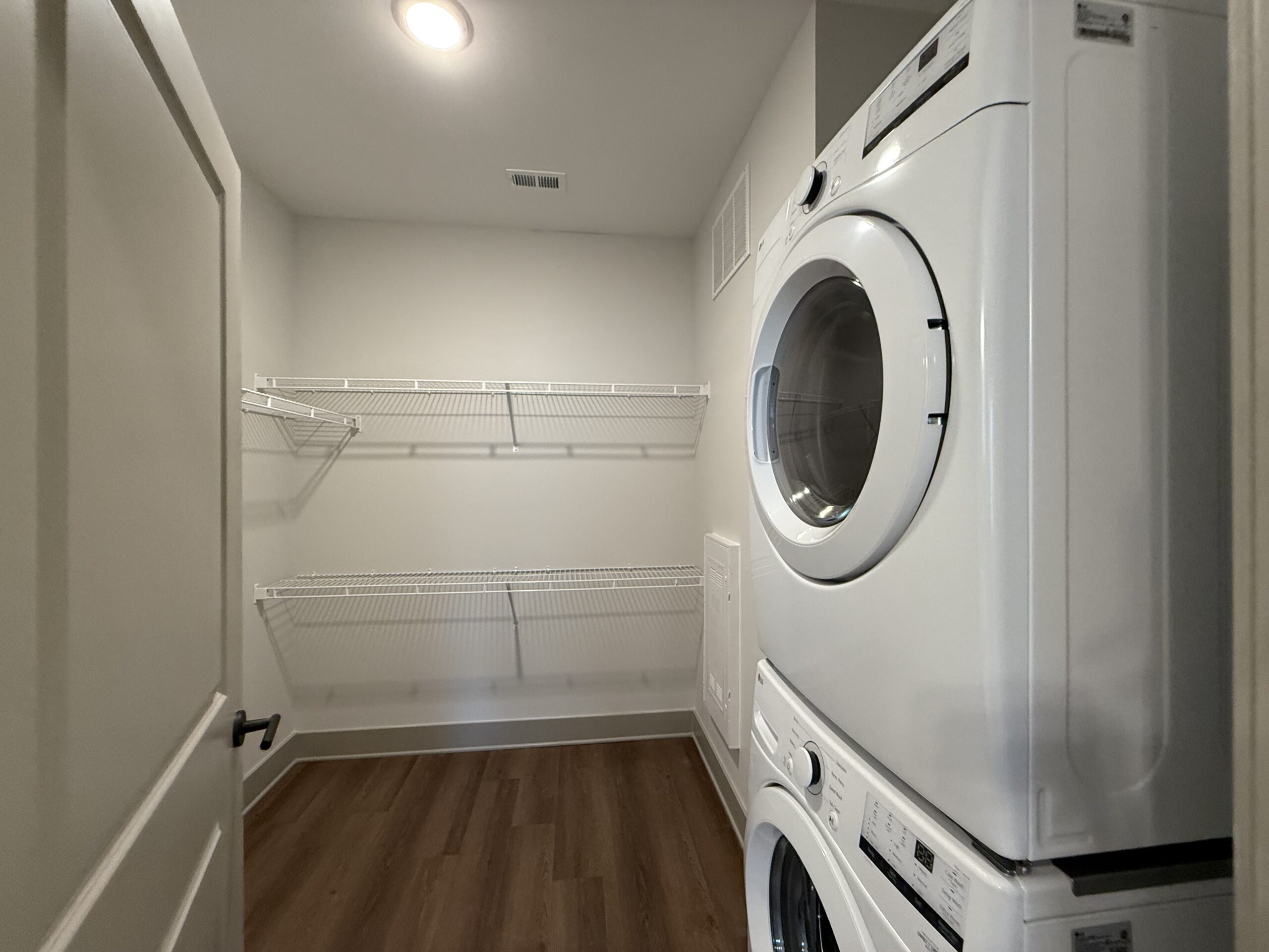 A laundry room features a stacked washer and dryer in white. The walls are painted a light color, and there are two wire shelving units mounted on the wall, providing storage space. The floor is made of wood, adding warmth to the space. A door is partially visible on the left side of the image.