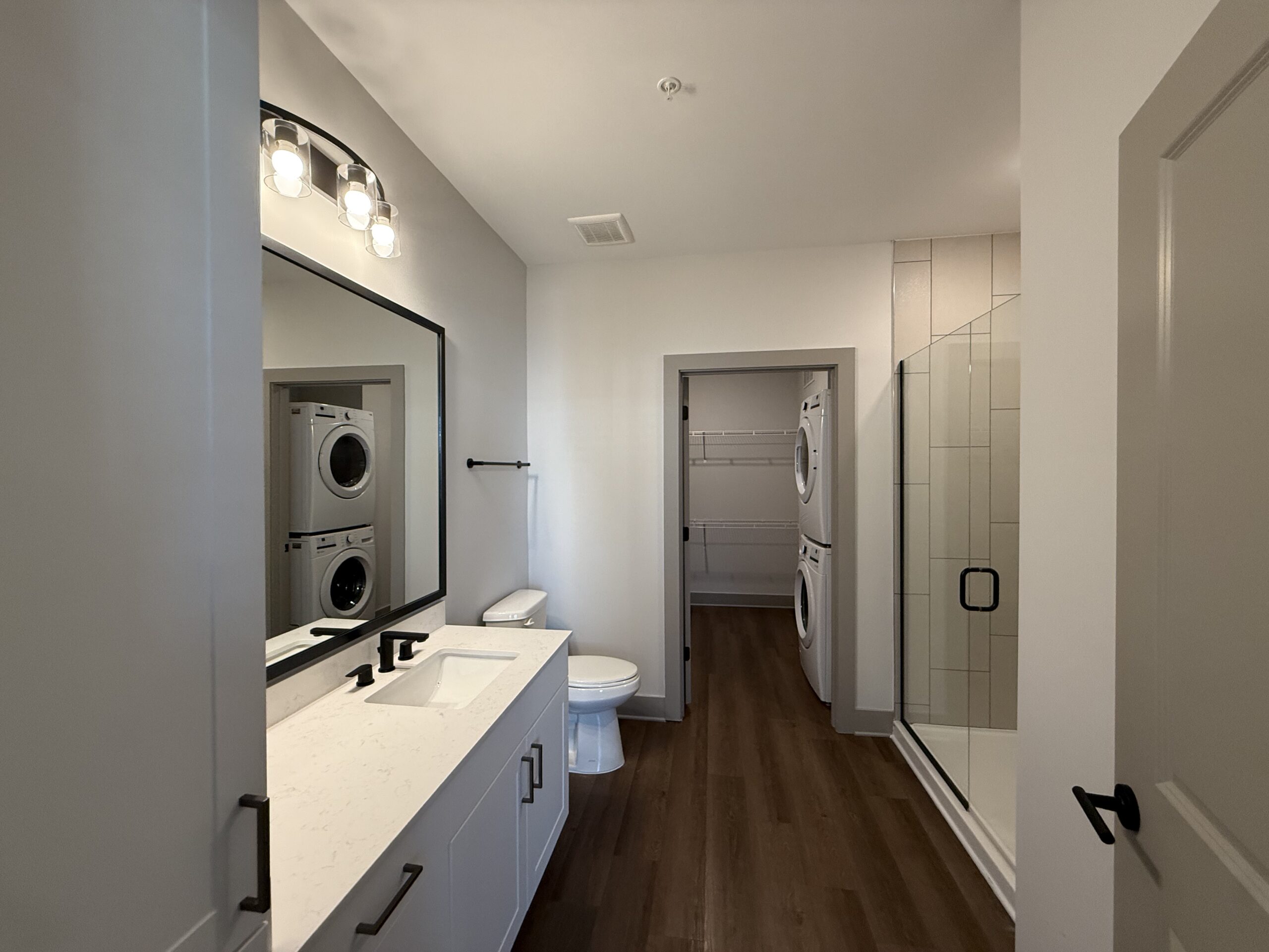 A modern bathroom features a double sink vanity with a white countertop and black fixtures. Above the vanity, a large rectangular mirror is framed in black. To the left, a toilet is positioned against the wall. A glass shower enclosure with tiled walls is visible on the right. In the background, a laundry area is seen with stacked washer and dryer units, and a small closet with wire shelving. The flooring is a warm wood tone, and the walls are painted in a light color, enhancing the spacious feel.