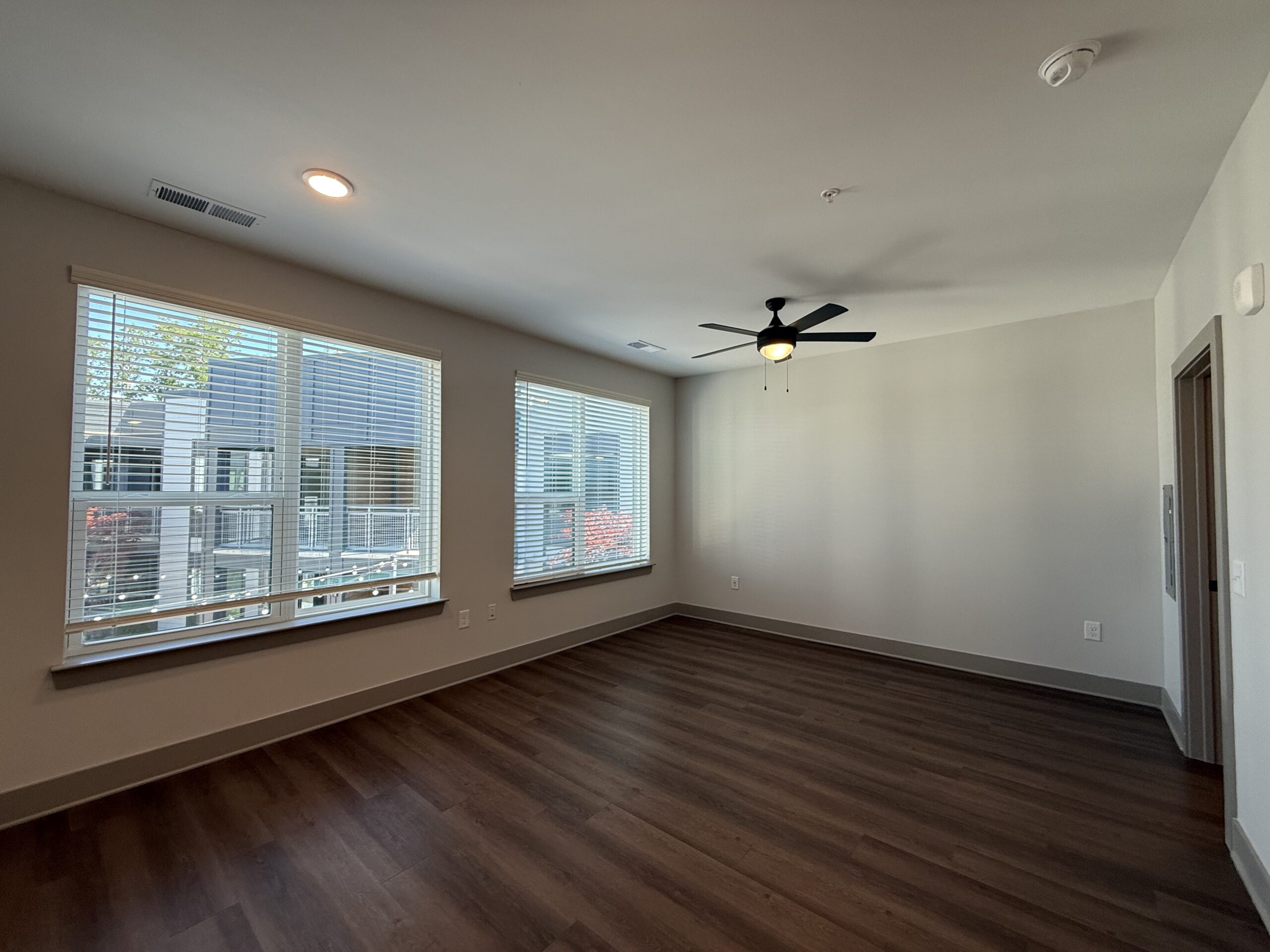 A spacious room features large windows with blinds, allowing natural light to fill the space. The walls are painted in a light color, and the flooring is a dark wood laminate. A ceiling fan with black blades hangs from the ceiling, providing a modern touch. In one corner, there is a door leading to another area, possibly a hallway or closet. The overall atmosphere is bright and inviting, suitable for various uses.