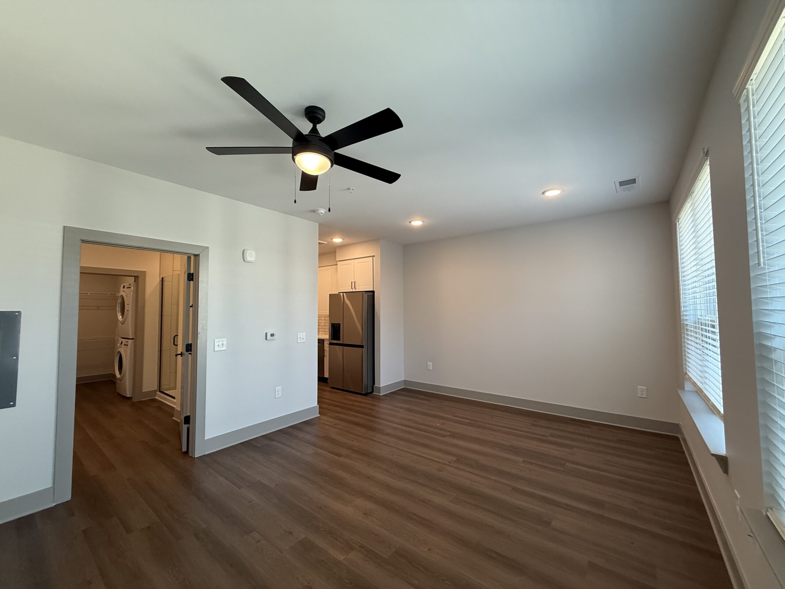 A modern living space featuring a ceiling fan with a light fixture. The room has light-colored walls and a wood-like floor. To the left, there is an open doorway leading to a laundry area with stacked washer and dryer units. In the background, a kitchen area is visible, showcasing a stainless steel refrigerator and white cabinetry. Natural light streams in through two windows with blinds on the right side of the room.