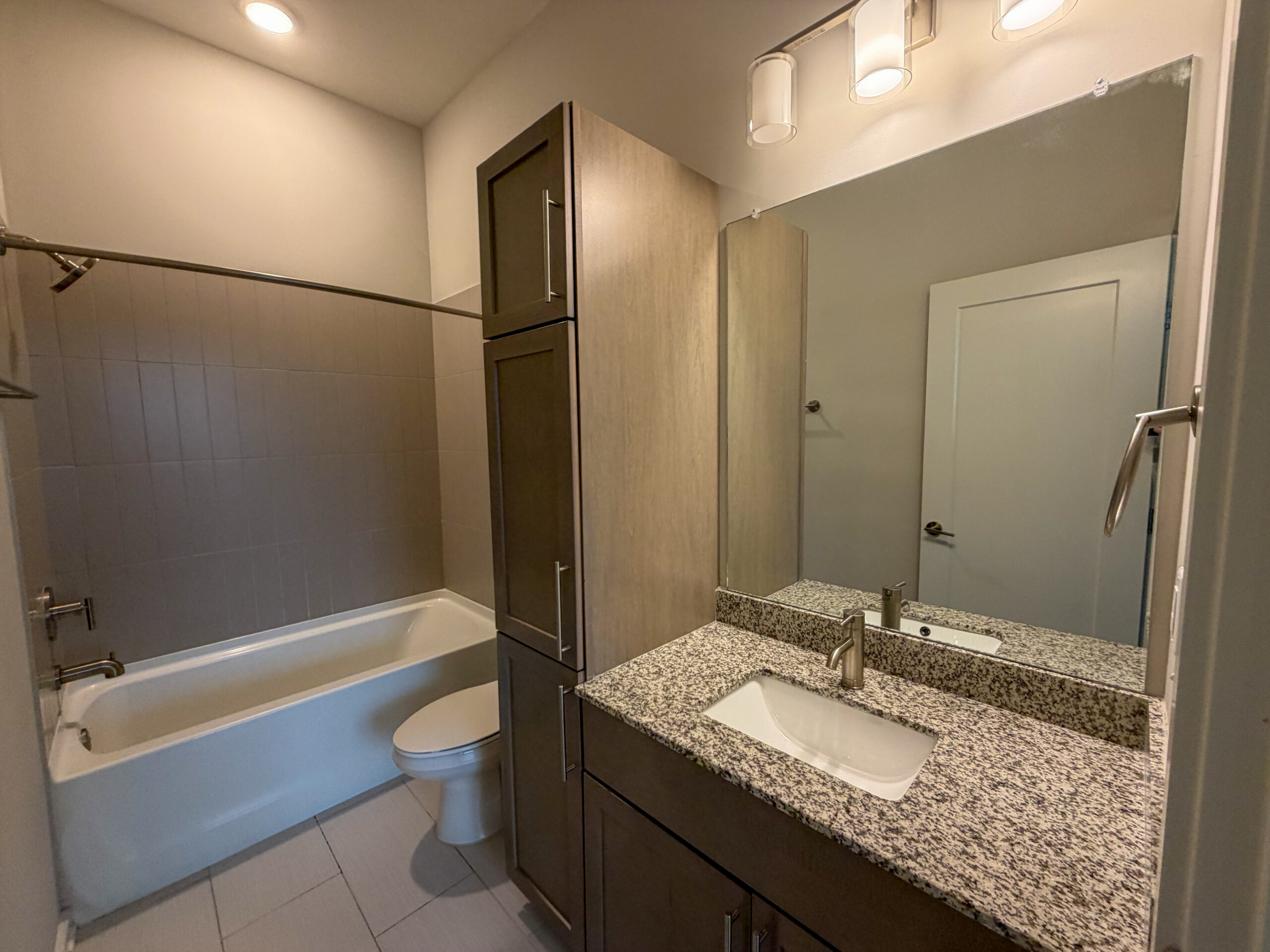 A modern bathroom featuring a white bathtub with a showerhead above, set against light gray tiled walls. To the right, there is a sleek vanity with a granite countertop and a rectangular sink. The cabinetry is a mix of dark and light wood tones, providing storage space. A large mirror is mounted above the sink, and a door is visible in the background, leading out of the bathroom. The overall design is clean and contemporary.