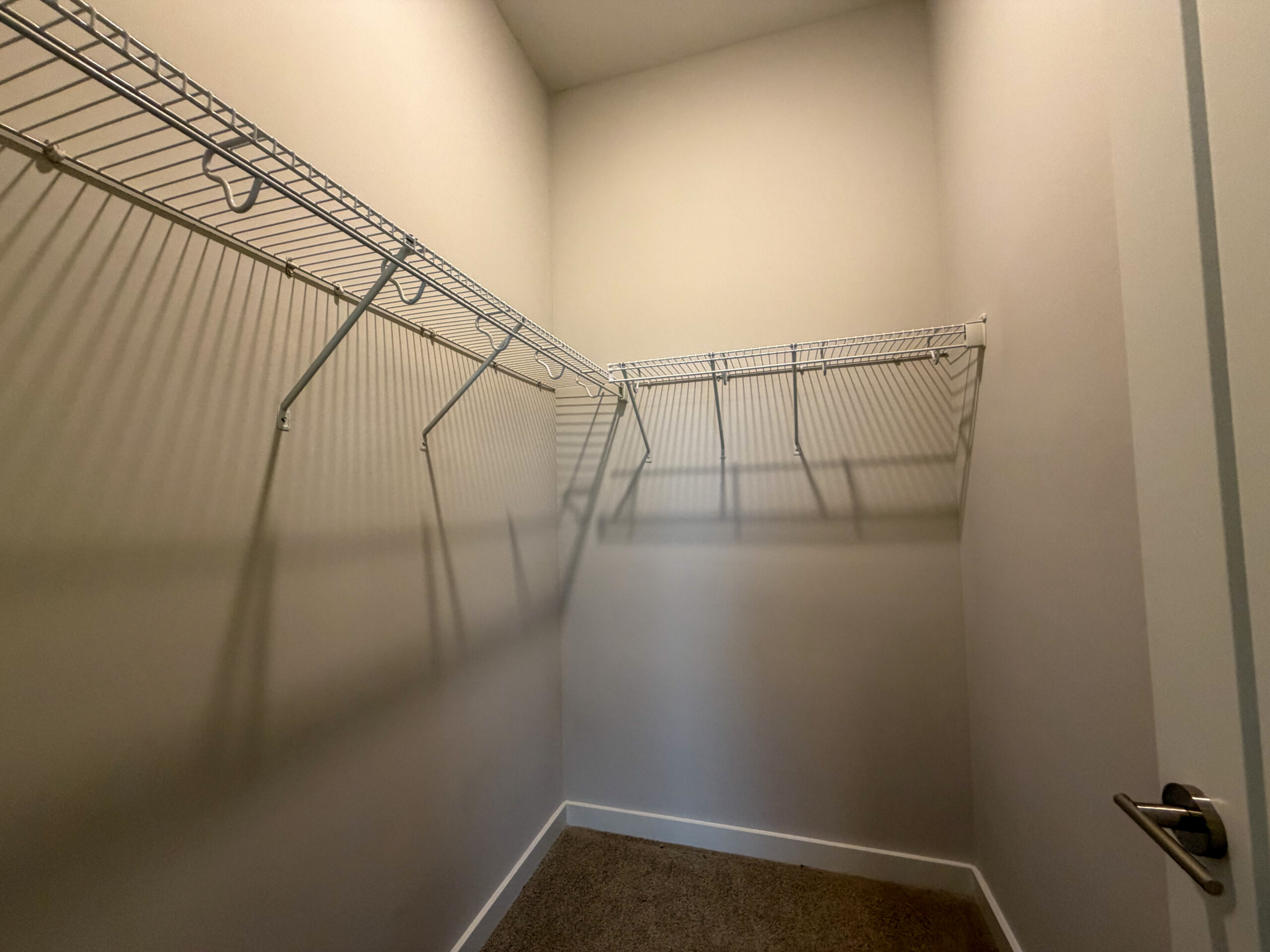 A small closet features two white wire shelving units mounted on the walls. The shelves are empty, casting shadows on the light-colored walls. The floor is bare, and a door handle is visible on the right side of the image. The overall space appears clean and organized, suitable for storage.