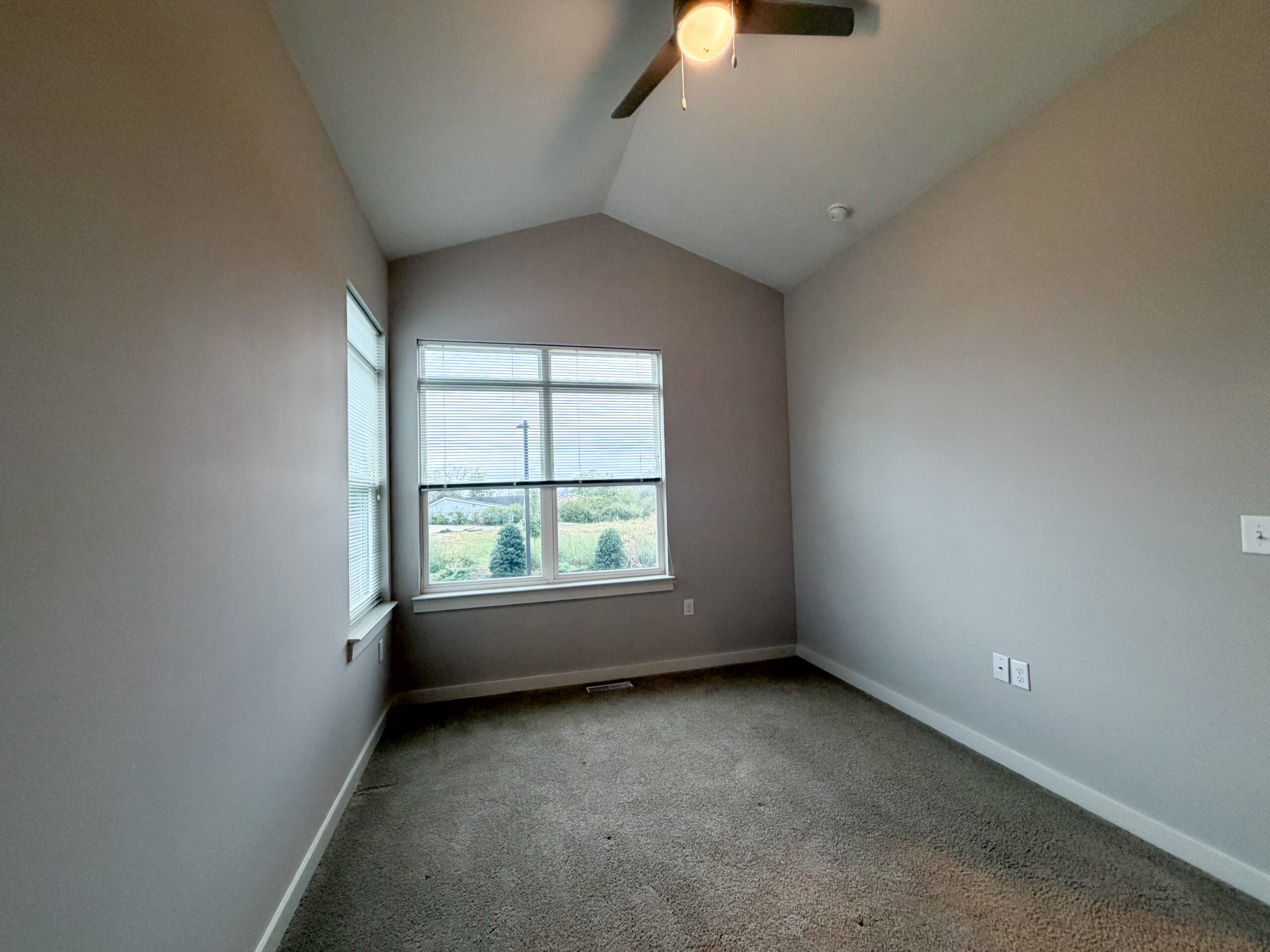 A small room with a high, sloped ceiling and light gray walls. There are two windows: one large window with blinds and a smaller window beside it. The floor is covered in beige carpet. The room appears empty, with electrical outlets visible on one wall. Outside the window, greenery and small trees are visible.