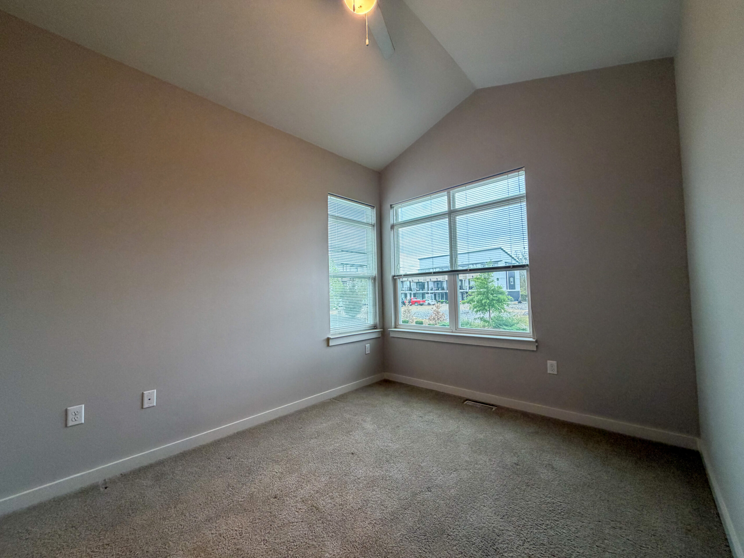 A spacious room with light gray walls and a high ceiling. Two large windows with blinds allow natural light to enter, offering a view of greenery and nearby buildings. The floor is covered with light-colored carpet, and there are electrical outlets on one wall. The overall atmosphere is bright and inviting.