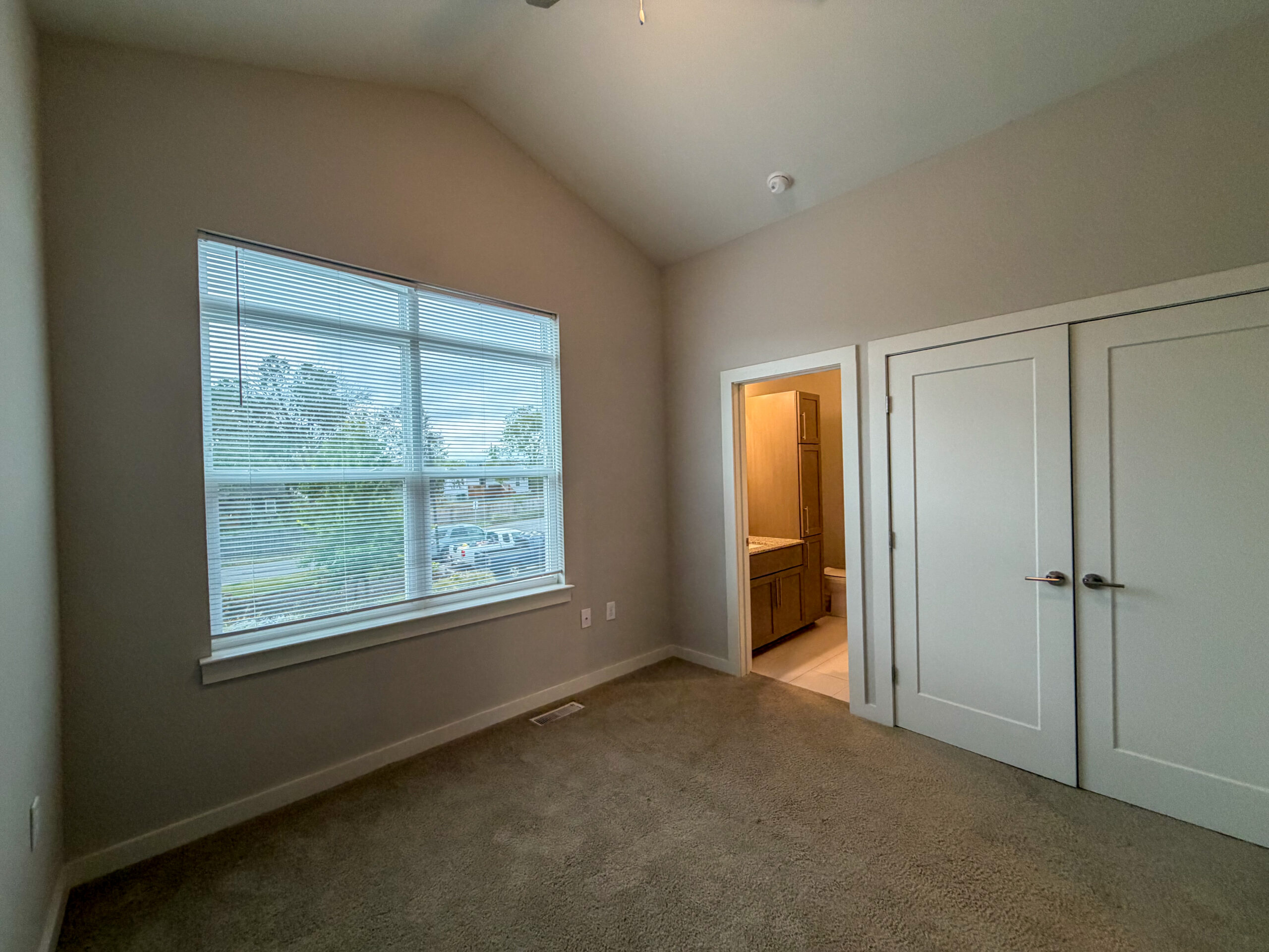 A well-lit room features a large window with blinds, allowing natural light to enter. The walls are painted in a soft, neutral color, and the floor is covered with light brown carpet. To the right, there is an open doorway leading to a bathroom area, which includes cabinetry and a toilet, visible through the entrance. The room has a high ceiling, contributing to a spacious feel.