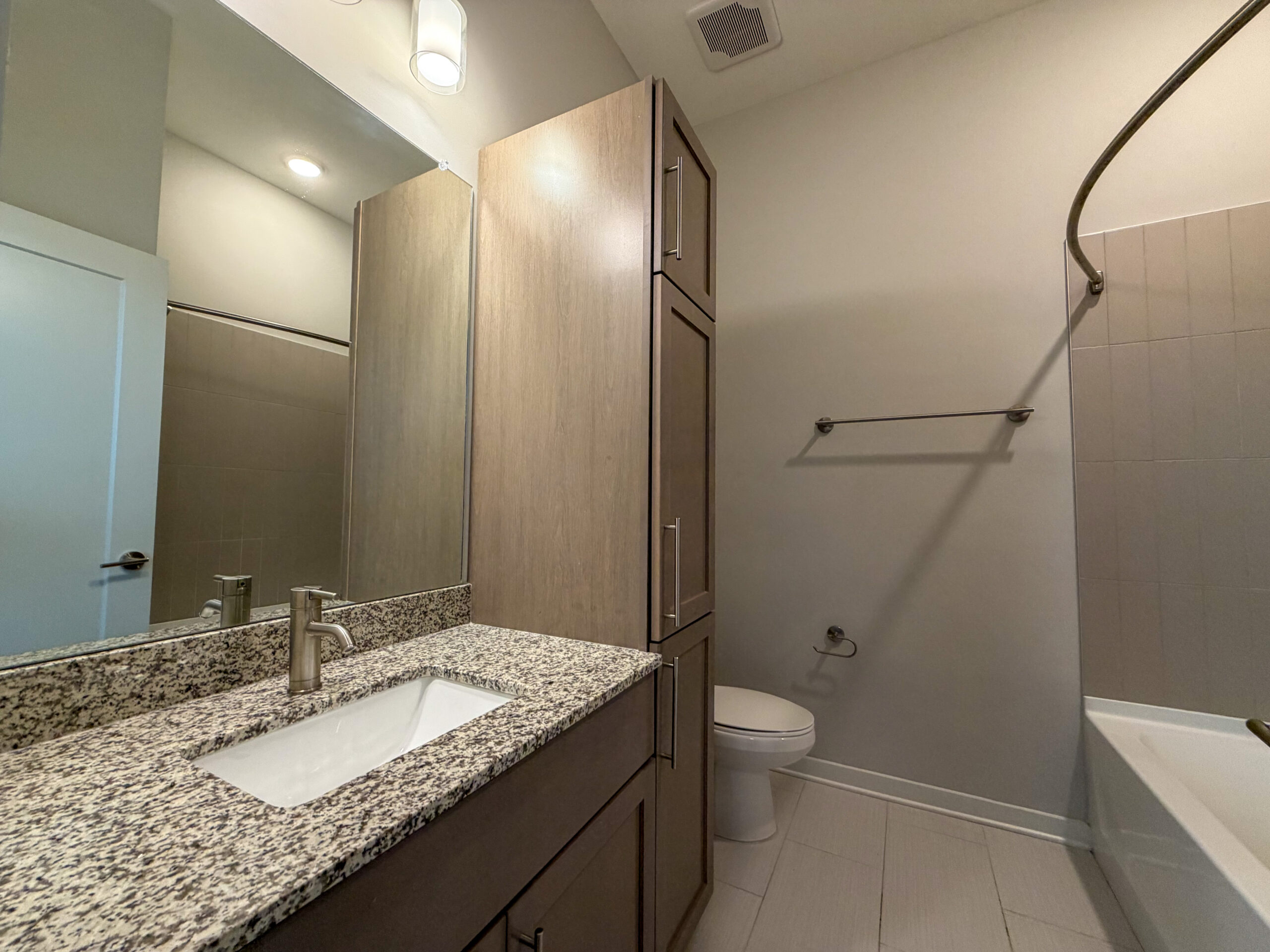 A modern bathroom featuring a granite countertop with an integrated sink and a sleek faucet. The cabinetry is light wood with minimalist handles. A large mirror reflects the space, enhancing the brightness. To the right, there is a shower area with a curved shower rod and a bathtub. A towel bar is mounted on the wall, and a white toilet is positioned nearby. The walls are painted in a neutral tone, and the flooring is tiled.