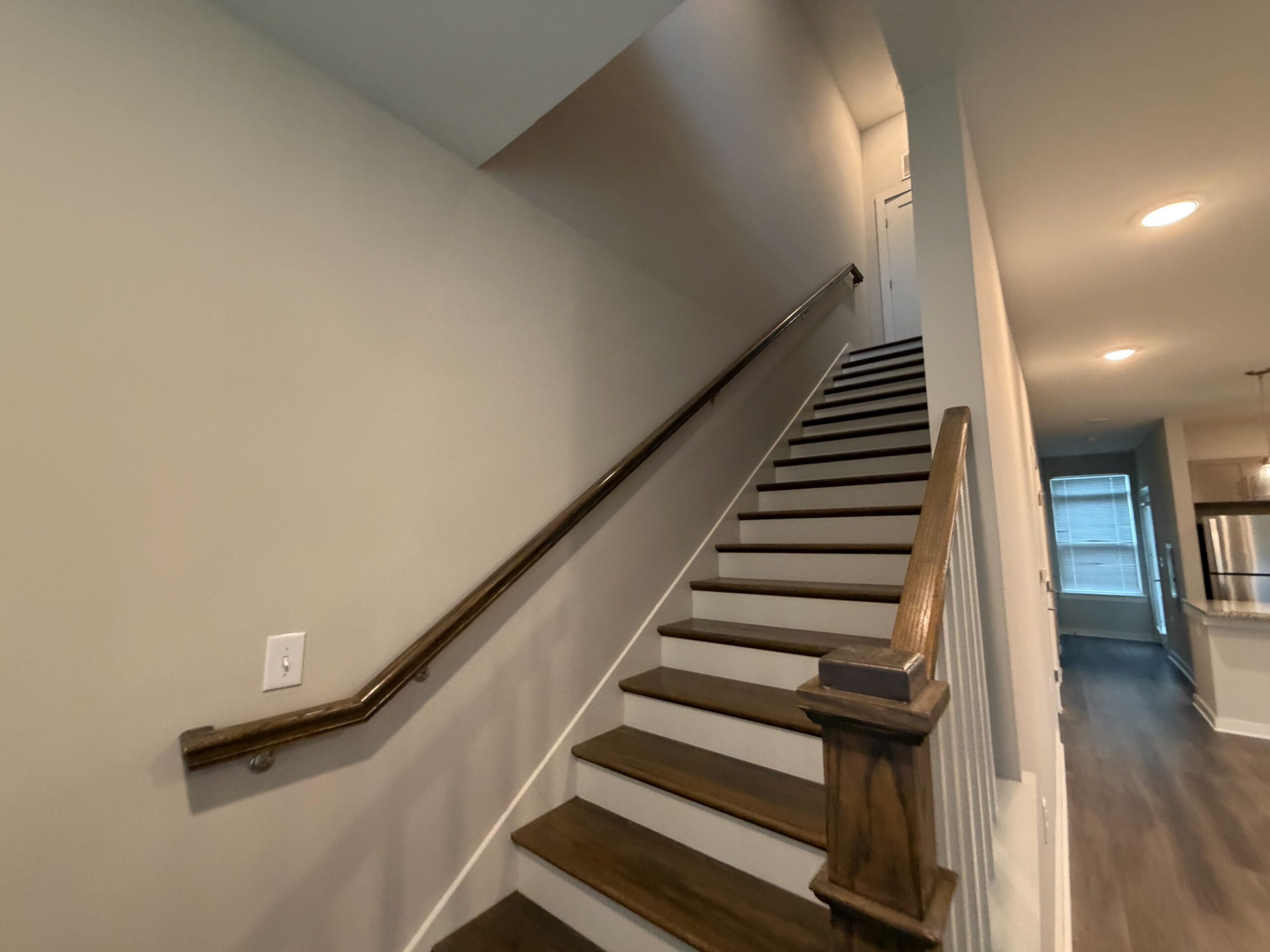 A staircase with wooden steps and a polished handrail ascends to an upper level. The walls are painted in a light neutral color, and the space is well-lit with recessed lighting. At the top of the stairs, a door is visible, leading to another room. To the right, a hallway extends towards a kitchen area, which features stainless steel appliances and a window with blinds. The flooring is a dark wood, adding warmth to the overall aesthetic.