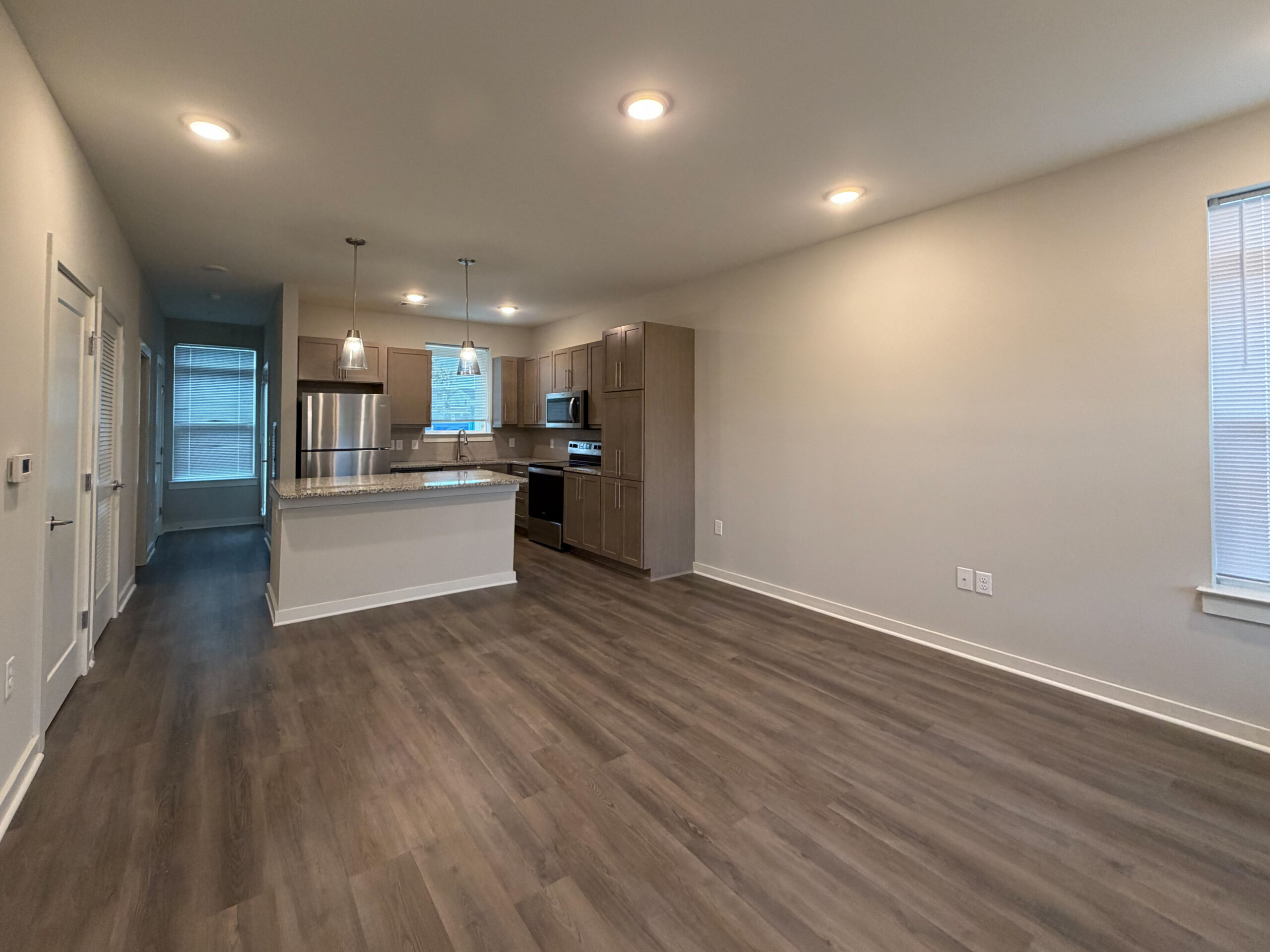 A modern, open-concept living space features a kitchen with light wood cabinetry, stainless steel appliances, and a granite countertop with a breakfast bar. Pendant lights hang above the bar area. The flooring is a dark laminate, and the walls are painted a light neutral color. A large window allows natural light to fill the room, and there are doors leading to other areas, including a hallway with additional light from a window. The overall atmosphere is bright and inviting.