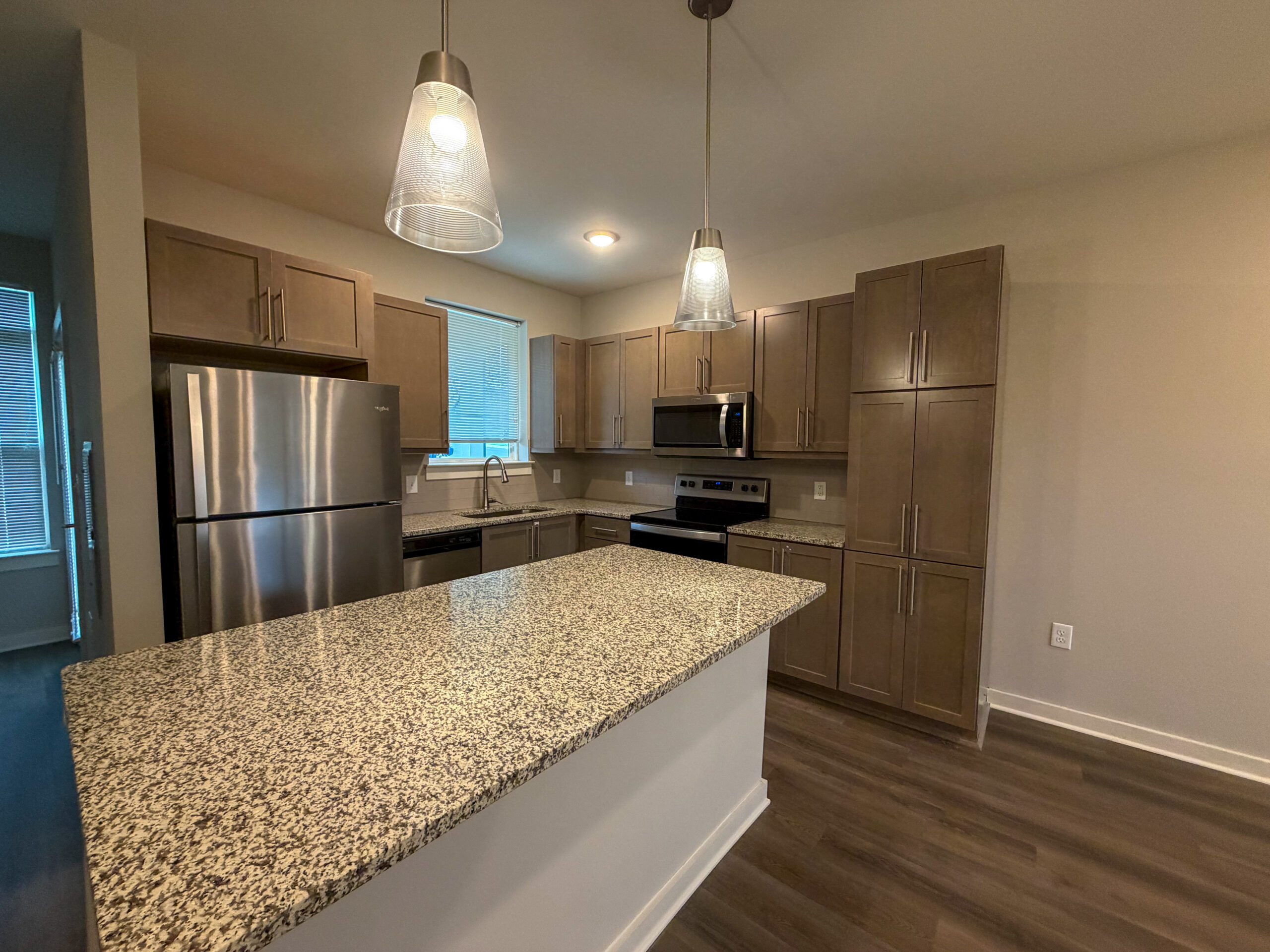 A modern kitchen featuring light brown cabinetry and a large granite countertop island. The kitchen is equipped with stainless steel appliances, including a refrigerator, microwave, and stove. A sink is positioned beneath a window, allowing natural light to enter. Two pendant lights hang above the island, and the flooring is a dark wood finish. The overall design is sleek and contemporary, with a neutral color palette.