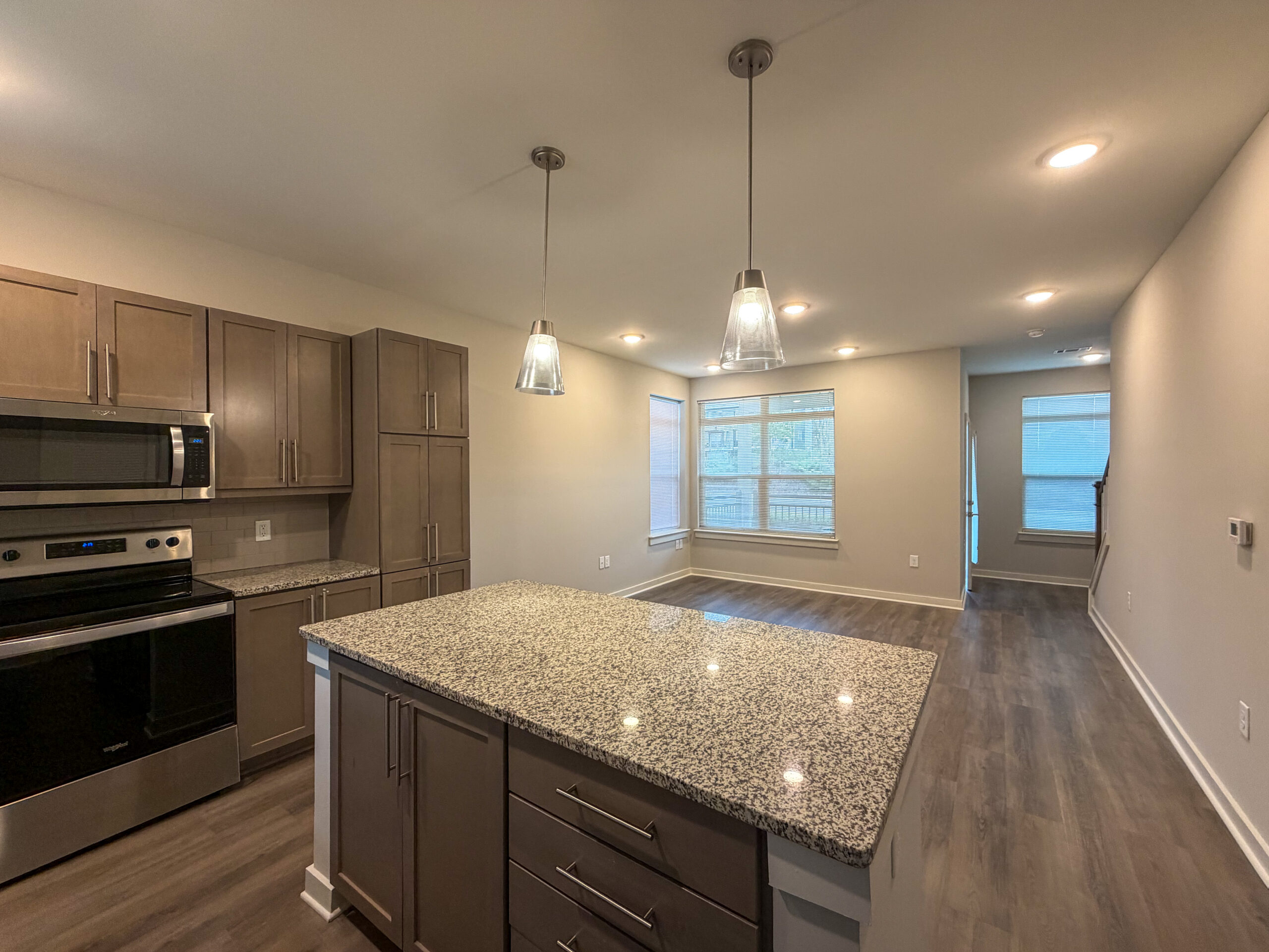 A modern kitchen features light brown cabinetry and a large granite countertop island. Stainless steel appliances, including a microwave and oven, are integrated into the cabinetry. Pendant lights hang above the island, providing illumination. The space opens into a living area with large windows allowing natural light, and a light-colored laminate floor extends throughout. The walls are painted in a neutral tone, enhancing the contemporary feel of the room.