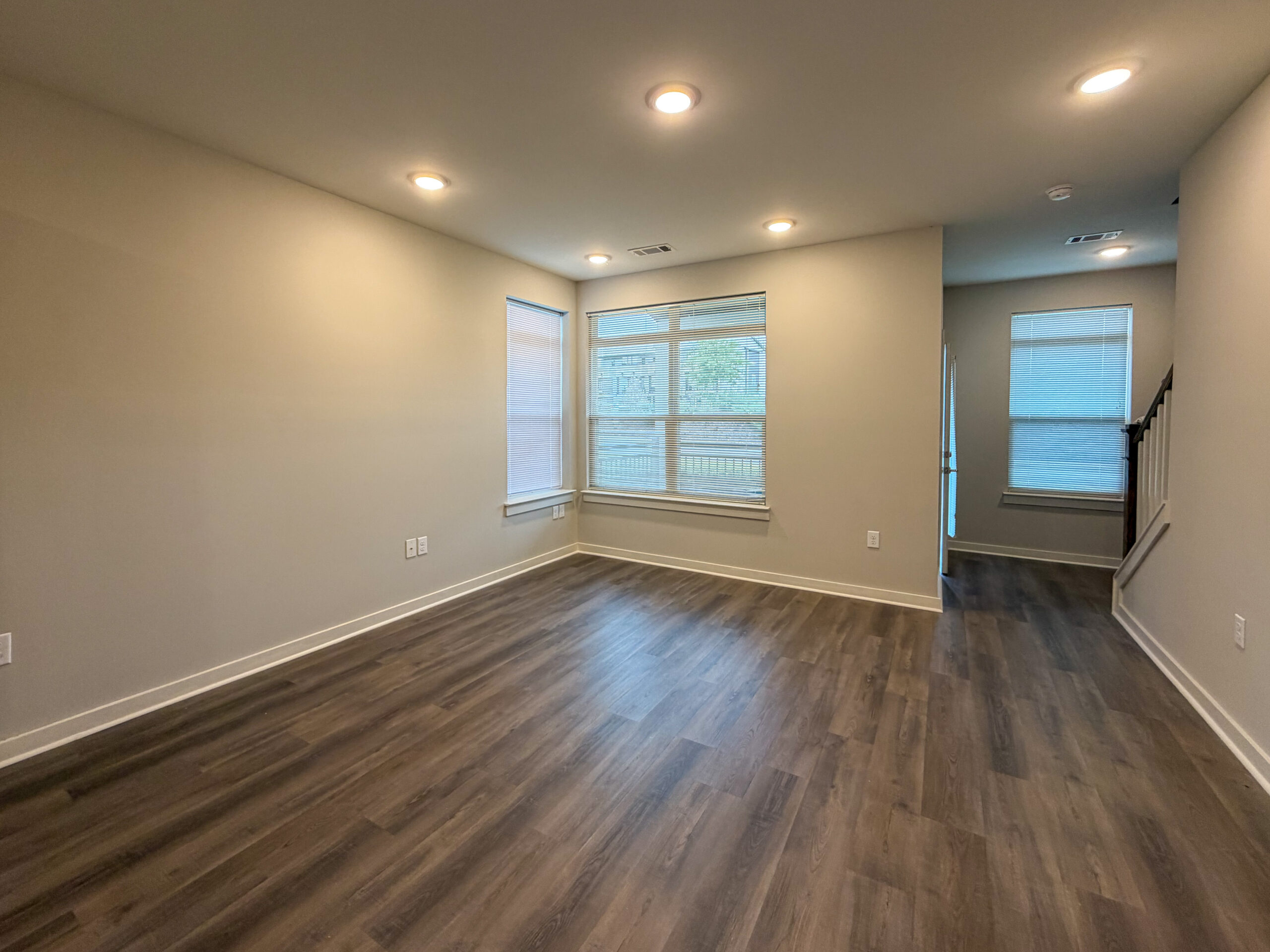 A spacious, well-lit room features light-colored walls and a modern laminate floor. Large windows with blinds allow natural light to fill the space, while recessed lighting adds additional illumination. In one corner, a staircase leads to an upper level, and a doorway on the right suggests access to another area. The overall ambiance is clean and inviting, suitable for various uses.