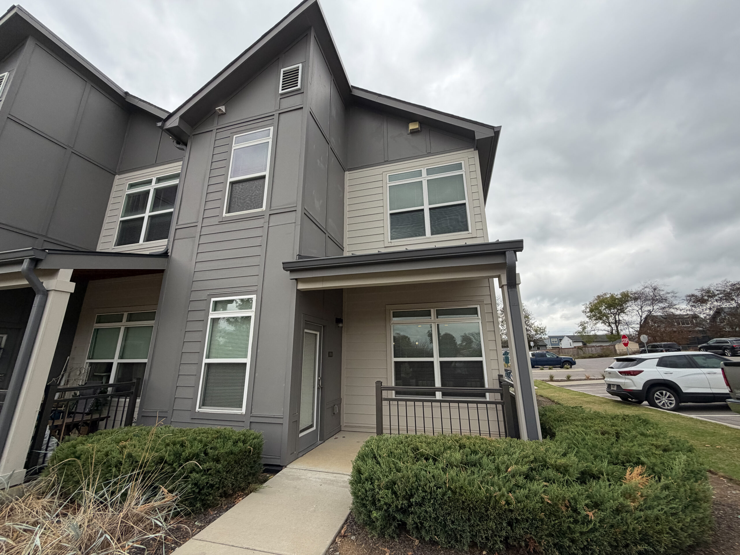 A modern two-story townhouse features a combination of gray siding and light-colored panels. The entrance has a small covered porch with a railing, and there are large windows on both levels, allowing natural light to enter. The landscaping includes neatly trimmed bushes in front of the porch. In the background, a parking area is visible with several vehicles and a stop sign. The sky is overcast, suggesting a cloudy day.