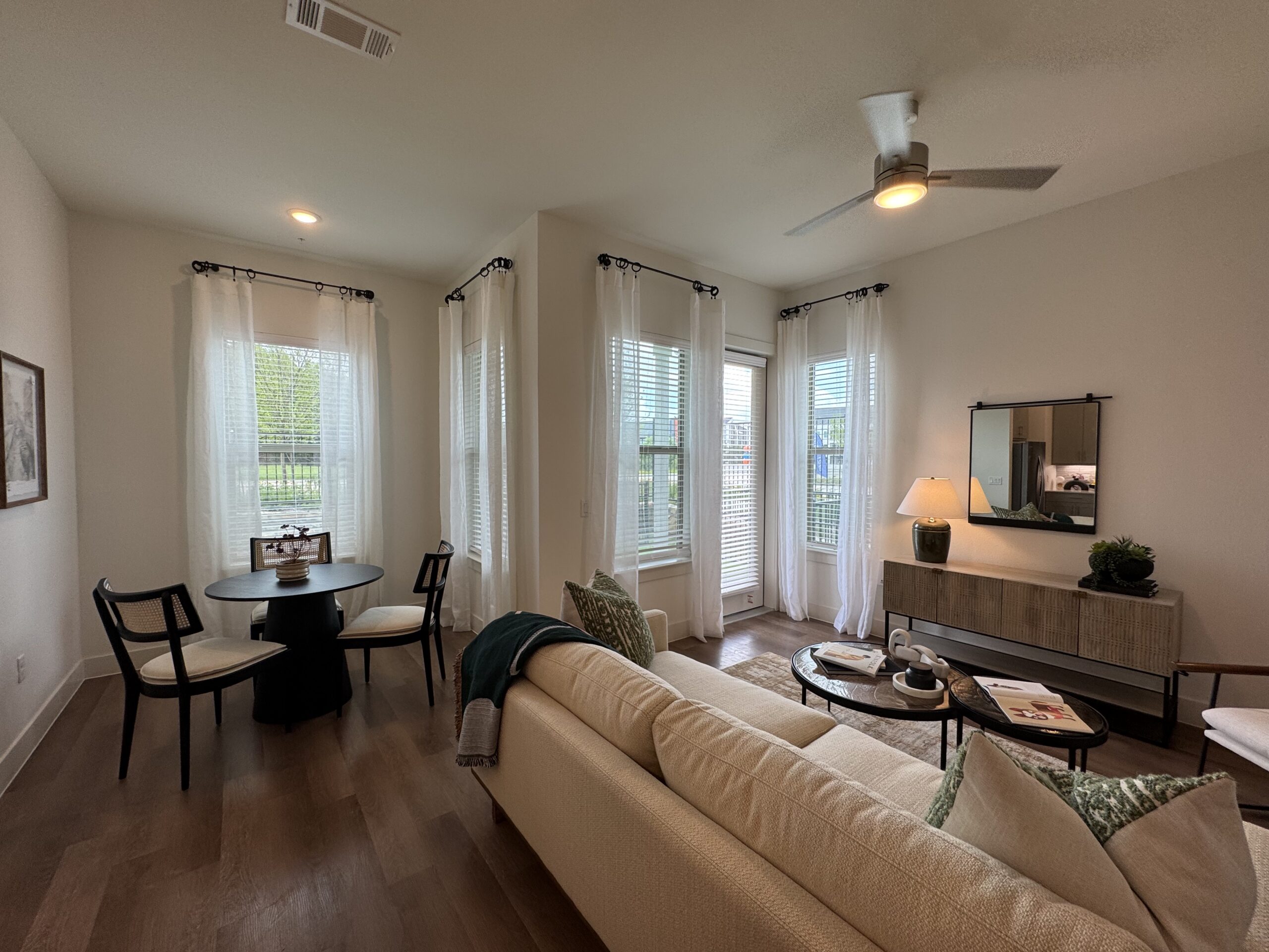 A cozy living space features a light-colored sofa adorned with decorative pillows. In the corner, a round black dining table is accompanied by three chairs, two of which have cushioned seats. Large windows with sheer white curtains allow natural light to fill the room, showcasing a view of greenery outside. A stylish sideboard with a lamp and decorative items sits against the wall, while a coffee table holds magazines and a decorative piece. The flooring is a warm wood tone, enhancing the inviting atmosphere.