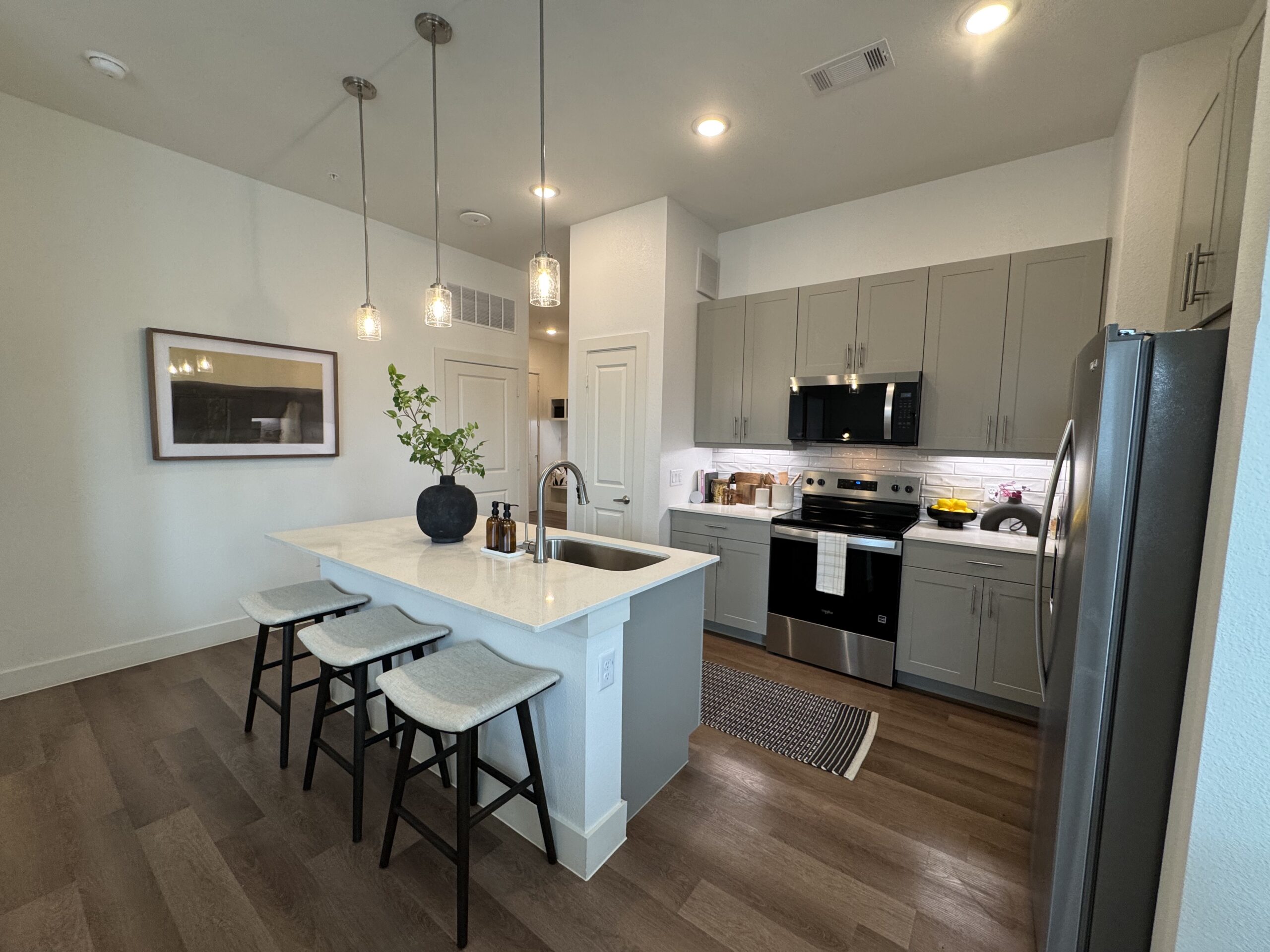 A modern kitchen features light gray cabinetry, a stainless steel refrigerator, and a black stove with a microwave above. The countertop is white with a subtle pattern, and there are three bar stools with light gray cushions. Above the island, three pendant lights provide illumination. A black vase with greenery sits on the counter, alongside decorative bottles. The floor is a warm wood tone, and a striped rug adds texture near the stove. A framed artwork hangs on the wall, complementing the contemporary design.