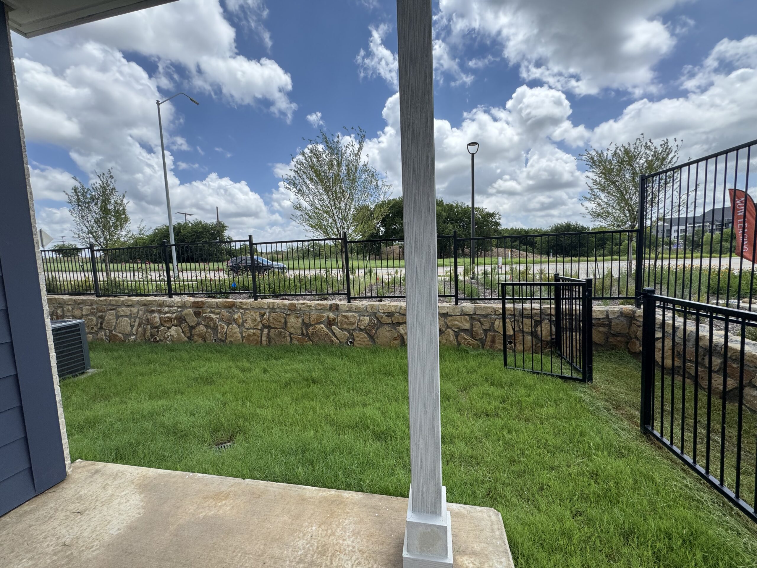 A grassy area is visible, bordered by a stone wall and a black metal fence. In the background, there are trees and a road with a few vehicles. The sky is partly cloudy, with a mix of blue and white clouds. A small gate in the fence is open, leading to the area beyond the stone wall.