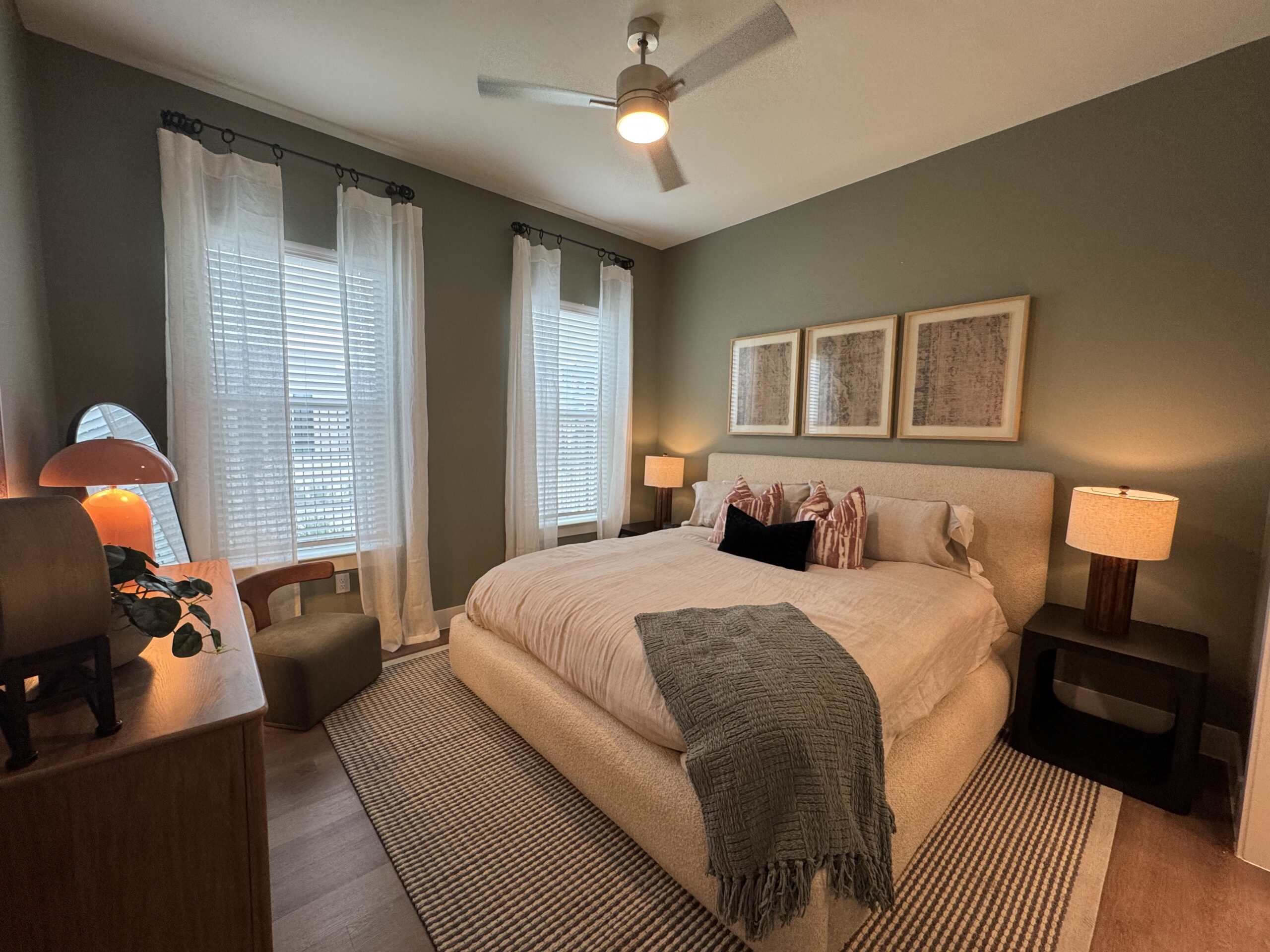 A cozy bedroom features a large bed with a light-colored, textured headboard and soft bedding. Decorative pillows in shades of pink and black add a pop of color. To the side, a wooden nightstand holds a warm-toned lamp. Natural light filters through sheer white curtains over a window with blinds. A stylish mirror and a small green accent chair complement the room's decor, which is set against a muted green wall. A patterned rug adds texture to the floor.