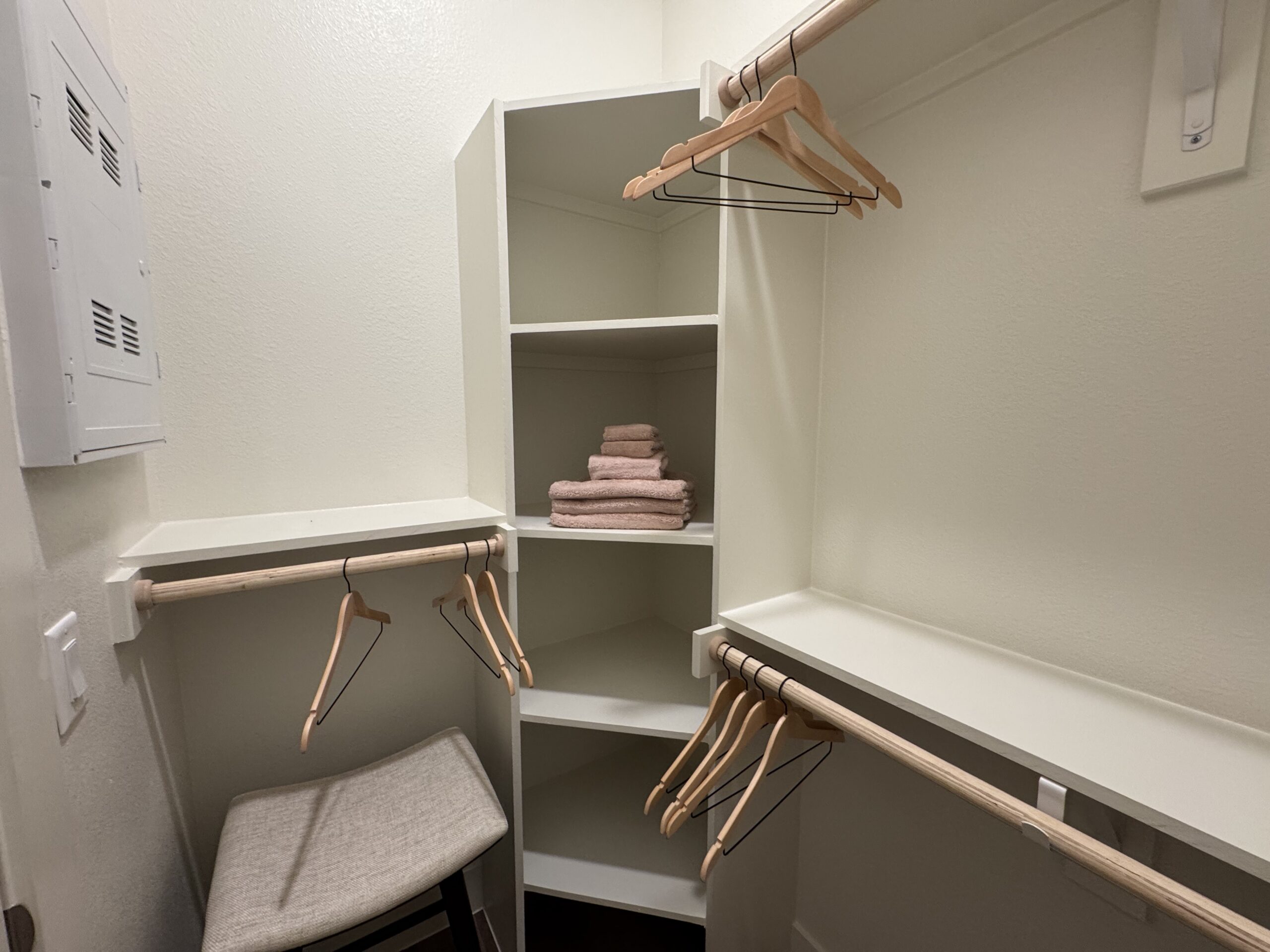 A small closet features white walls and shelving. On the left, there is a wooden hanging rod with several wooden hangers. A stack of soft, pink towels is neatly arranged on one of the shelves. The closet also includes an empty shelf and a small, cushioned stool or seat at the bottom. A white electrical panel is mounted on the wall.