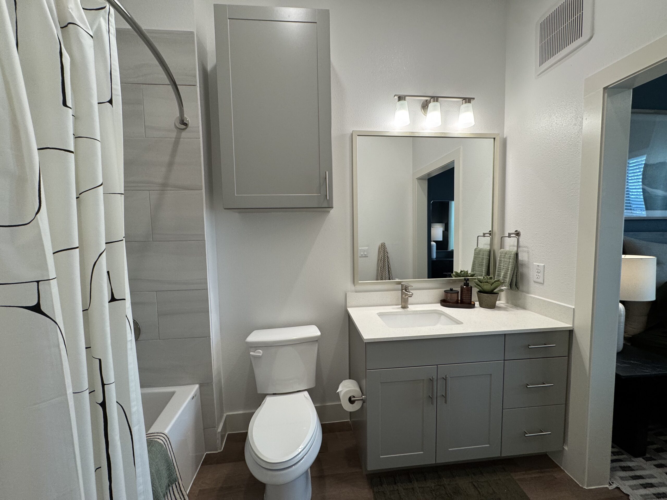 A modern bathroom features a light gray vanity with a white countertop and a square sink. Above the sink is a rectangular mirror framed in light gray, illuminated by three glass light fixtures. To the left, a shower area is partially obscured by a white shower curtain with black geometric lines. The wall behind the shower is tiled in light gray, and a bathtub is visible below the showerhead. A white toilet is positioned next to the vanity. Decorative elements include two green towels hanging on towel bars and a small potted plant on the countertop. The overall color scheme is neutral and contemporary.