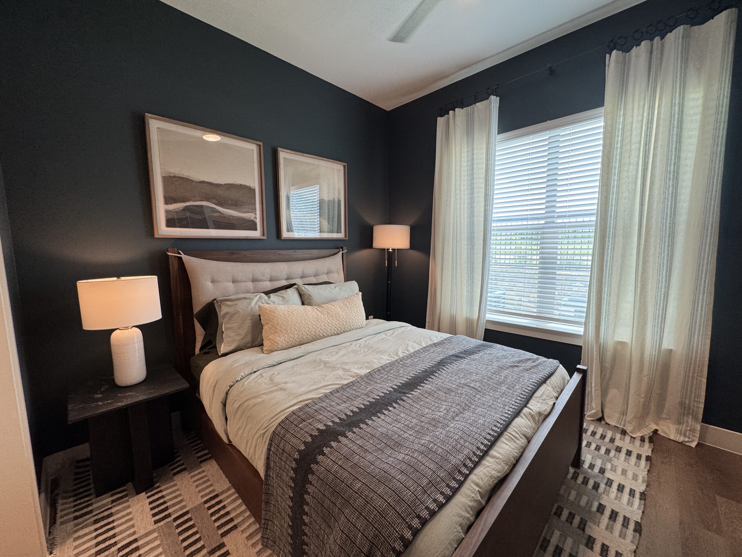 A cozy bedroom features a queen-sized bed with a tufted headboard, dressed in light-colored bedding and a textured throw blanket. Two framed landscape artworks hang above the bed against a dark green wall. A bedside table with a white lamp sits next to the bed, while a tall floor lamp provides additional lighting. Sheer, light-colored curtains frame a window with horizontal blinds, allowing natural light to filter in. The floor is covered with a patterned rug, adding warmth to the space.