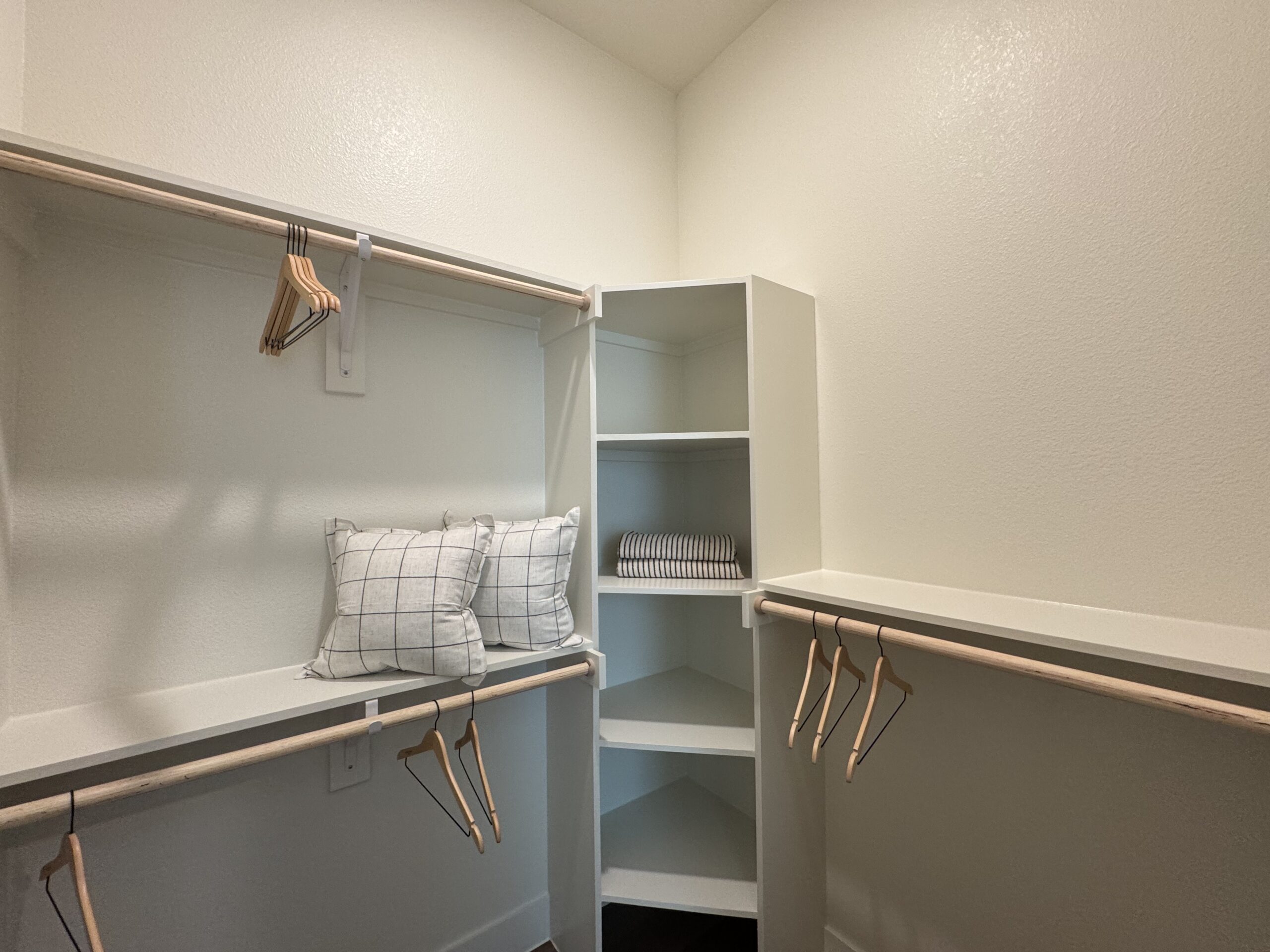 A well-organized closet features a light-colored interior with a wooden hanging rod and several wooden hangers. On the left side, there are two decorative pillows with a grid pattern resting on a shelf. To the right, a small shelving unit contains neatly folded striped towels. The overall design is clean and minimalist, with ample space for storage.