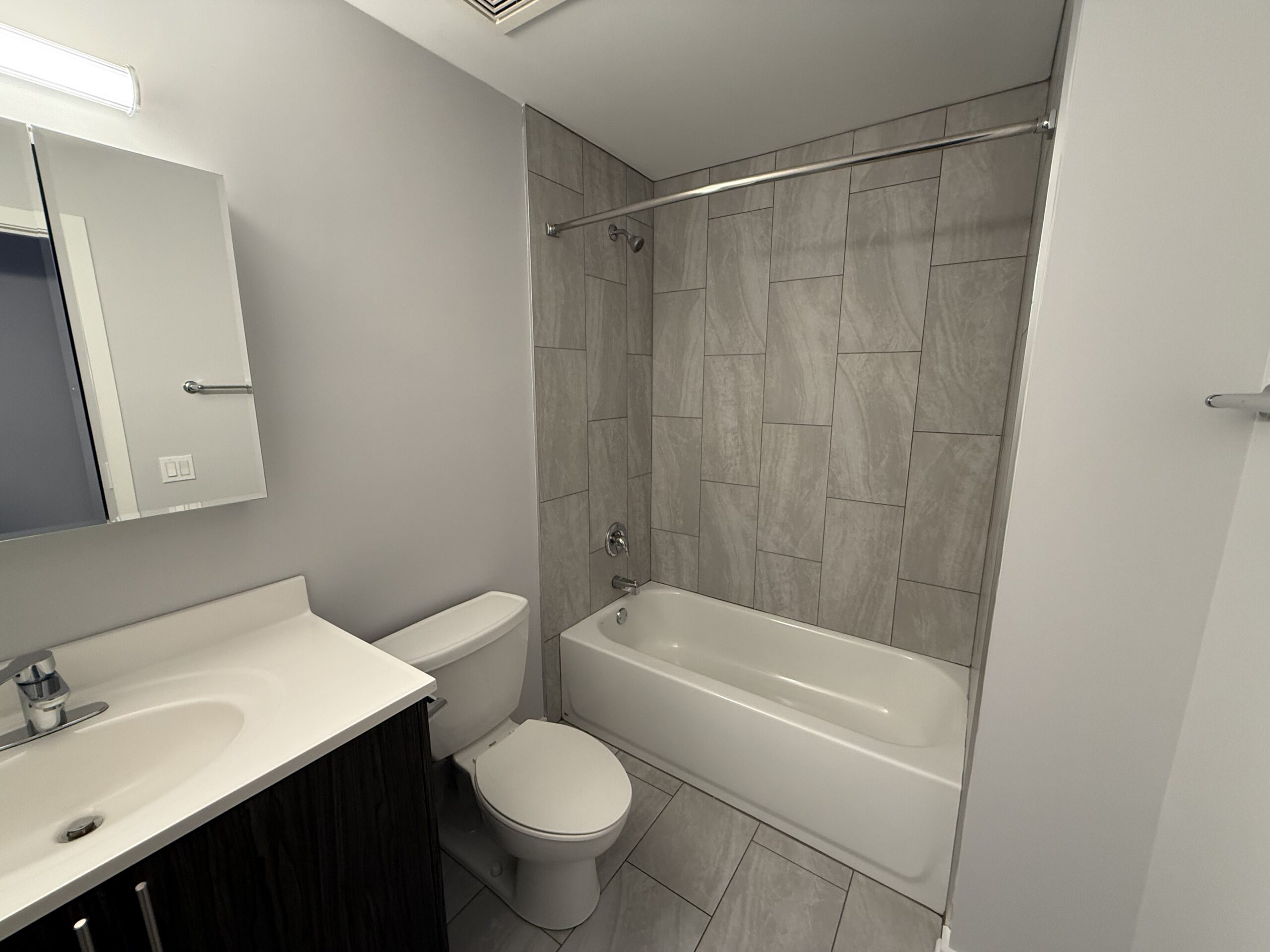 A modern bathroom features light gray tiled walls and a matching floor. On the left, there is a white vanity with a sink and a sleek faucet. Above the vanity, a rectangular mirror is mounted on the wall. To the right, a white toilet is positioned next to a bathtub with a showerhead above it. A chrome shower rod extends over the tub. The overall color scheme is neutral and contemporary.