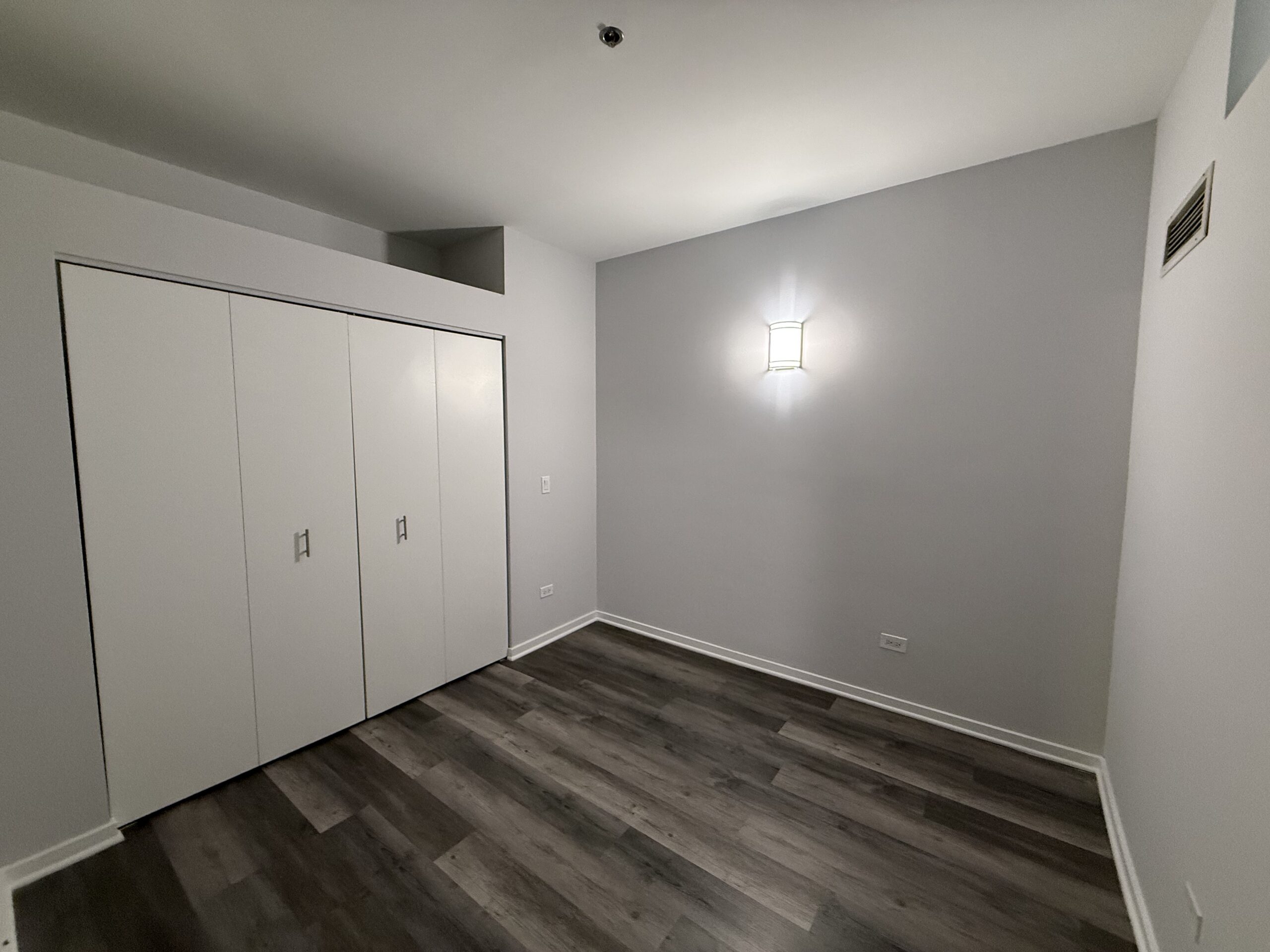 A small, minimally furnished room featuring light gray walls and a wood-like floor. On the left side, there is a set of white sliding closet doors with simple metal handles. A modern wall sconce provides soft lighting on the right wall. The room has a clean and uncluttered appearance, with a small vent visible near the ceiling.