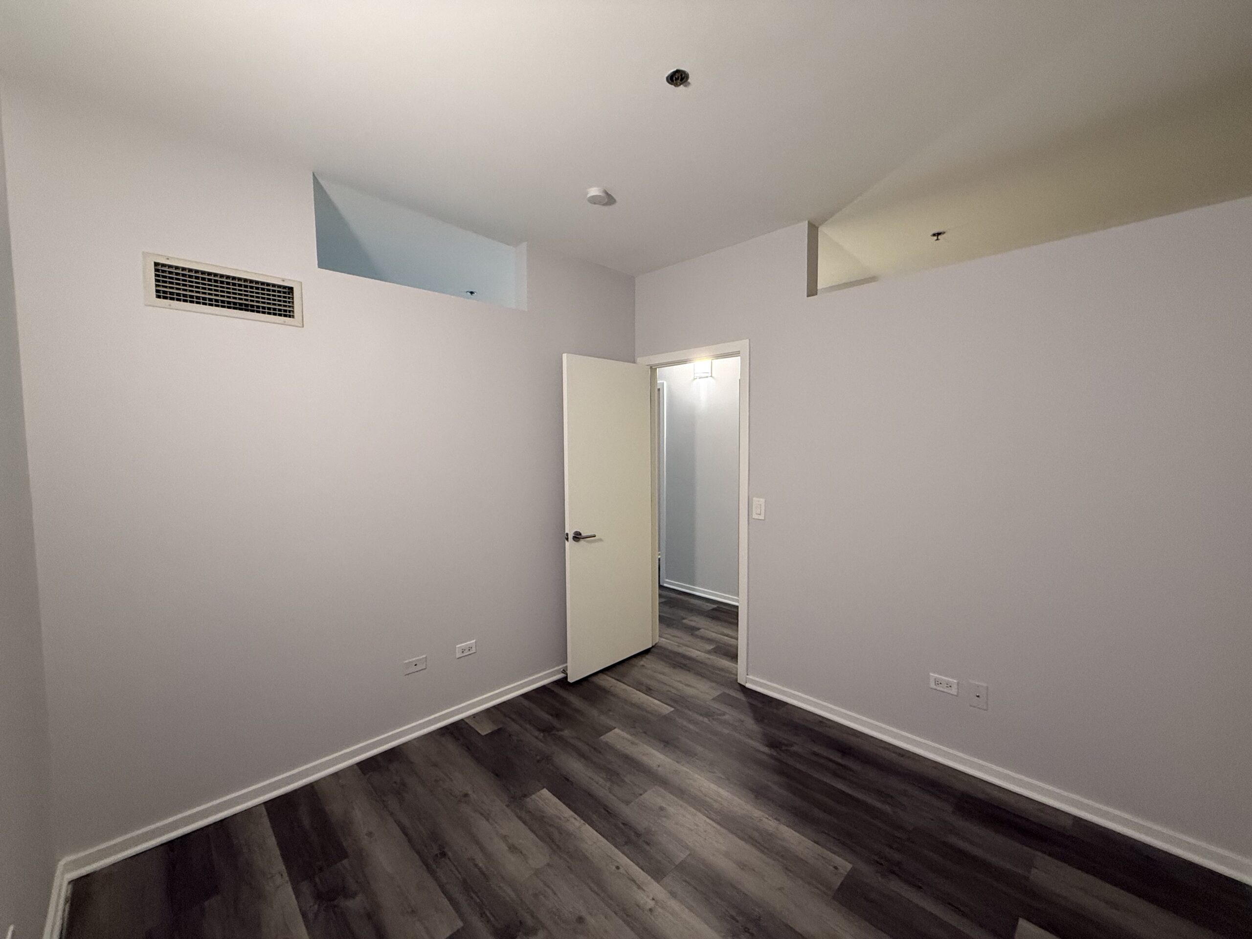A small, empty room with light gray walls and dark wood-like flooring. There is a white door partially open, leading to another space. Above the door, a small rectangular opening is visible, possibly for ventilation. A vent is mounted on the wall to the left, and a ceiling light fixture is present. The overall atmosphere is bright and minimalistic.