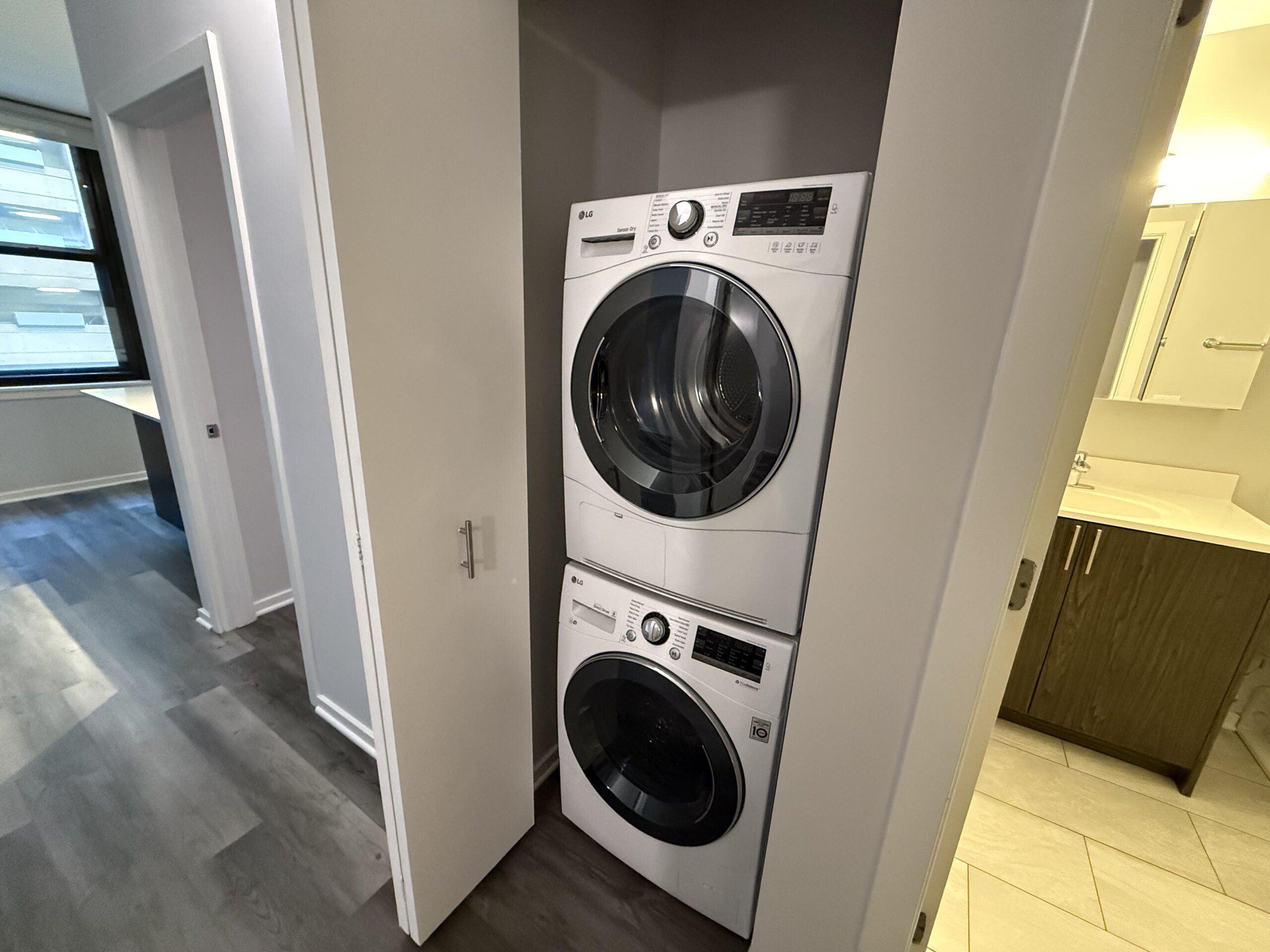 A stacked washer and dryer set in white is positioned against a wall. The dryer is on top, featuring a large circular door and a control panel with various settings. Below it, the washing machine has a similar design with a smaller door and control panel. The surrounding area includes a doorway leading to a bathroom with a light-colored countertop and dark cabinetry, and a window allowing natural light into the space. The flooring is a light gray laminate.