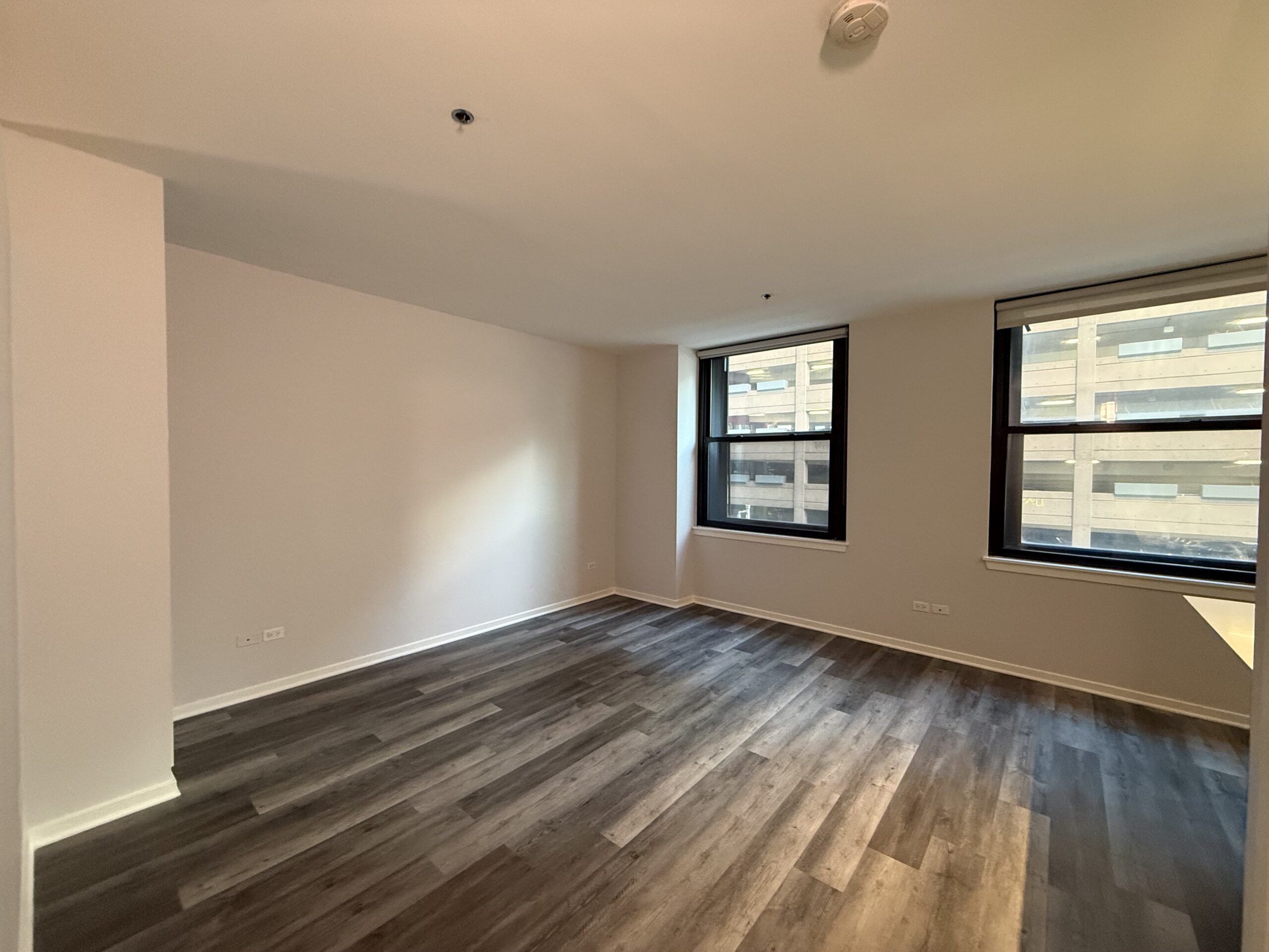 A spacious room with light-colored walls and dark wood-like flooring. Two large windows allow natural light to enter, revealing a view of a neighboring building. The room is empty, providing a blank canvas for potential furnishings. The ceiling is plain, and there are no visible light fixtures.