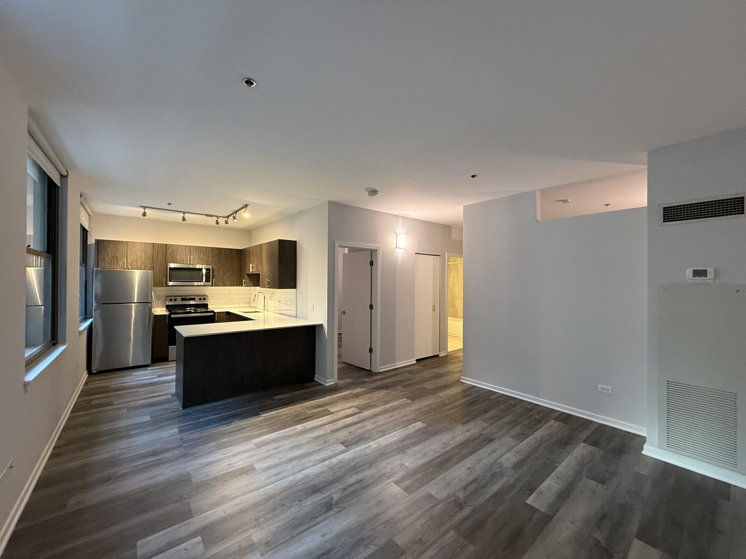 A modern kitchen and living area are visible, featuring dark wood cabinetry and stainless steel appliances, including a refrigerator and an oven. The kitchen island has a white countertop. The flooring is a light wood laminate. Natural light enters through large windows, and there are recessed lights in the ceiling. To the right, there are doors leading to a bathroom and additional rooms, with a light fixture illuminating the hallway. The walls are painted in a soft gray tone.