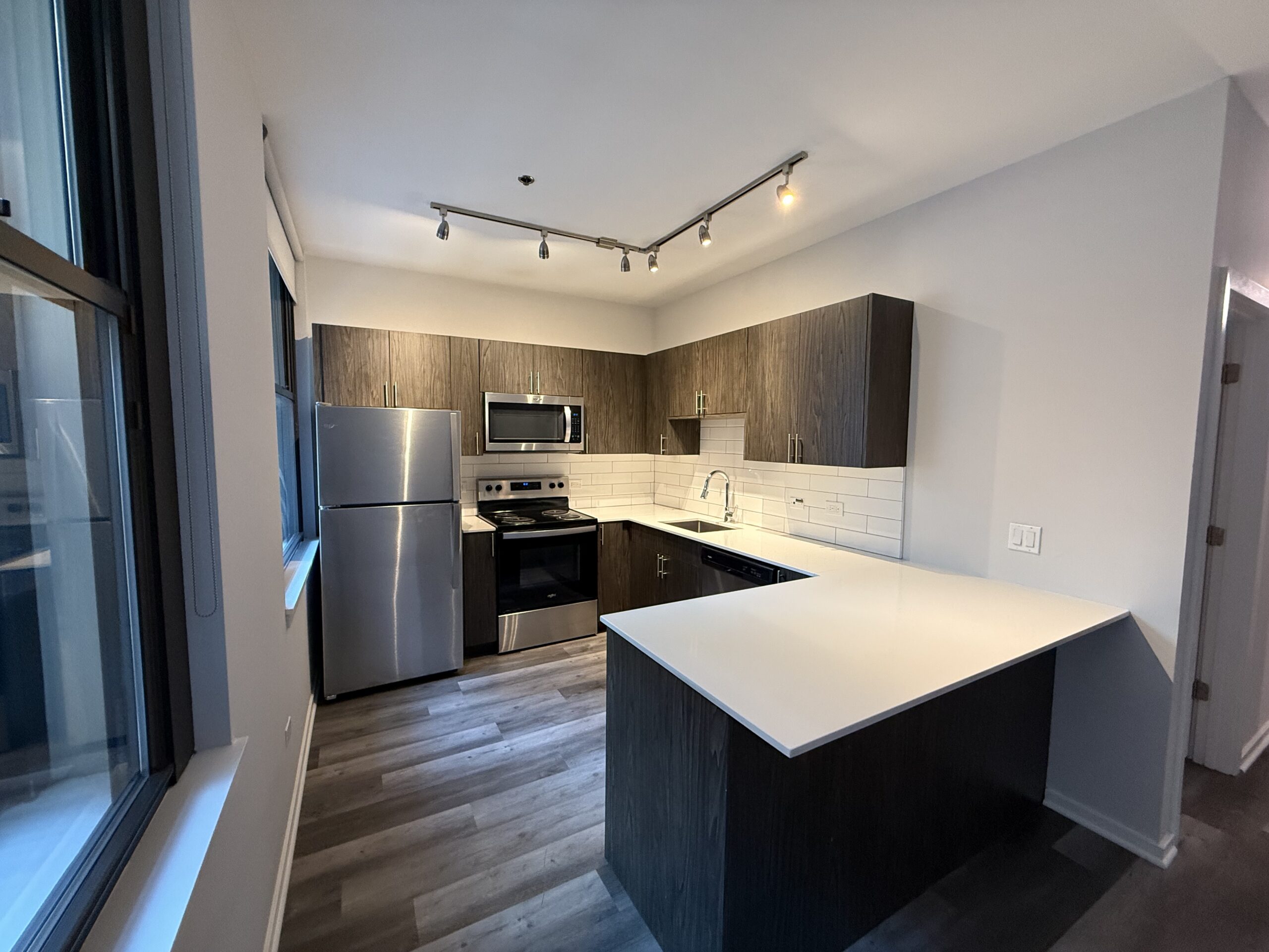 A modern kitchen featuring dark wood cabinetry and a white countertop. Stainless steel appliances include a refrigerator, microwave, and oven. The backsplash consists of white subway tiles, and there is a sleek sink with a faucet. The space is well-lit with track lighting and has large windows allowing natural light to enter. The flooring is a light wood laminate, enhancing the contemporary feel of the kitchen.