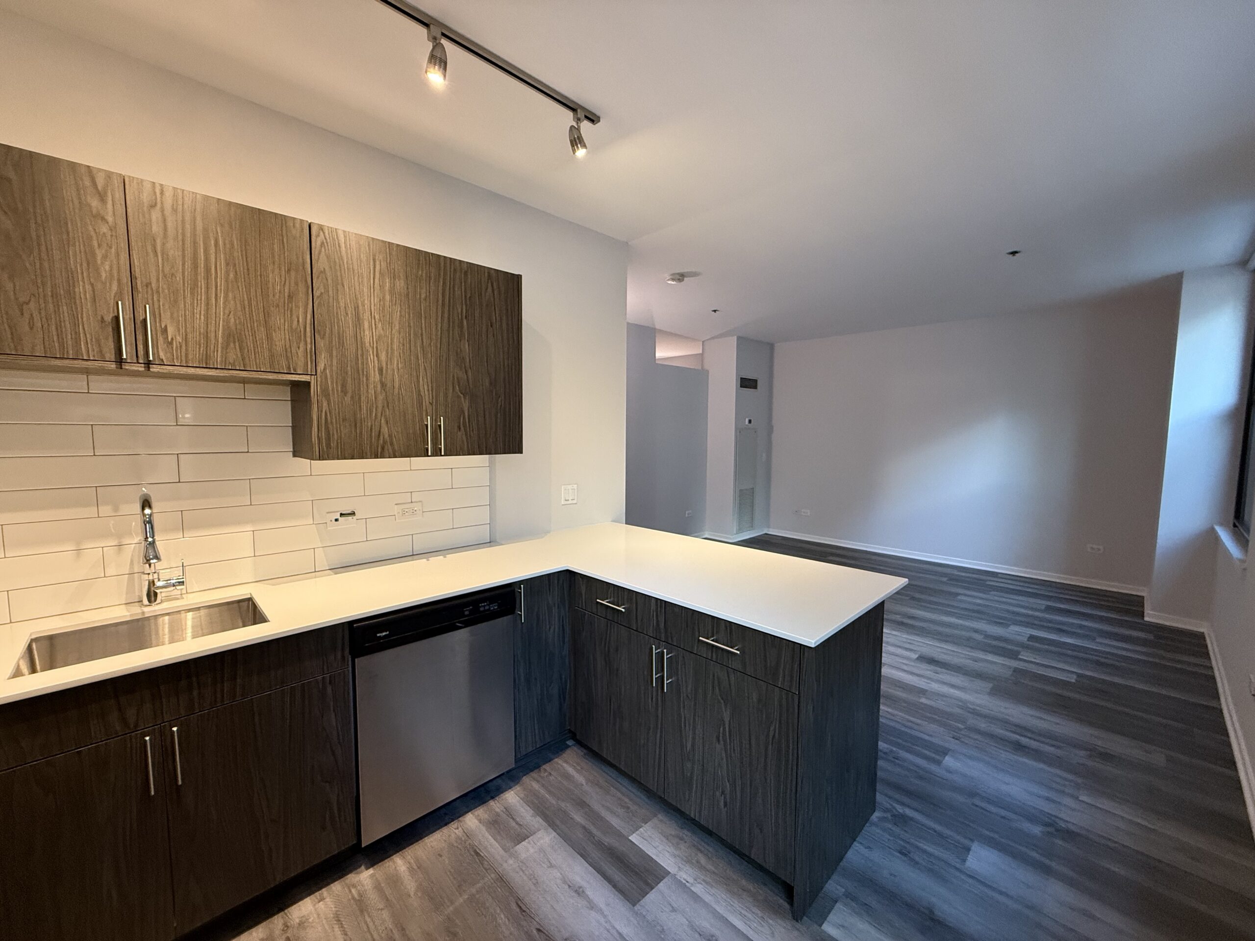 A modern kitchen features dark wood cabinetry with sleek handles, complemented by a white countertop and a stainless steel sink. The backsplash consists of white subway tiles. To the left, a stainless steel dishwasher is visible. The flooring is a light gray wood-like laminate, and the space opens up to a well-lit living area with neutral walls and a window allowing natural light.