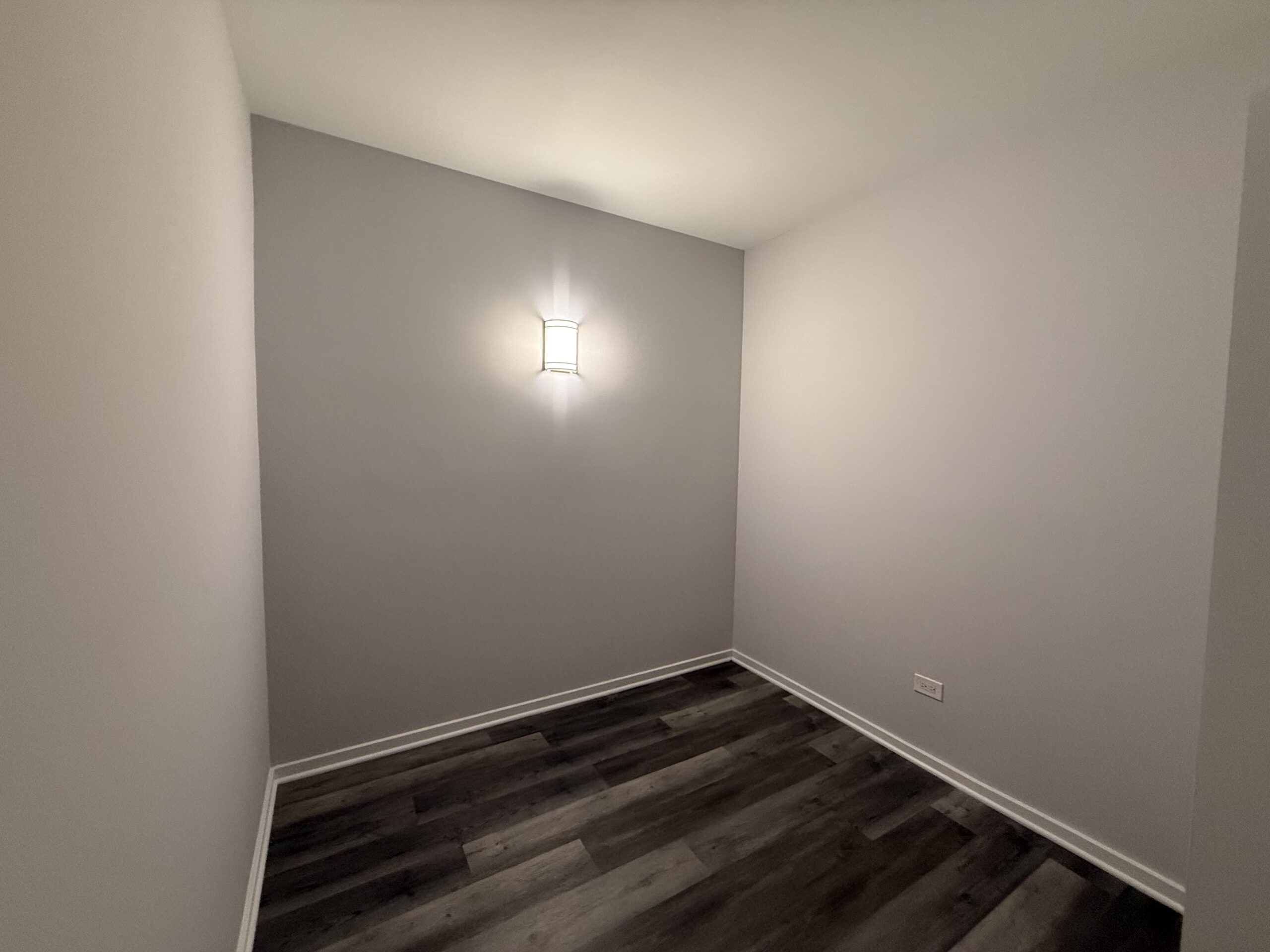 A small, empty room with light gray walls and a dark wood laminate floor. A modern wall sconce is mounted on one wall, providing soft illumination. The room has no furniture, creating a minimalist and open feel.