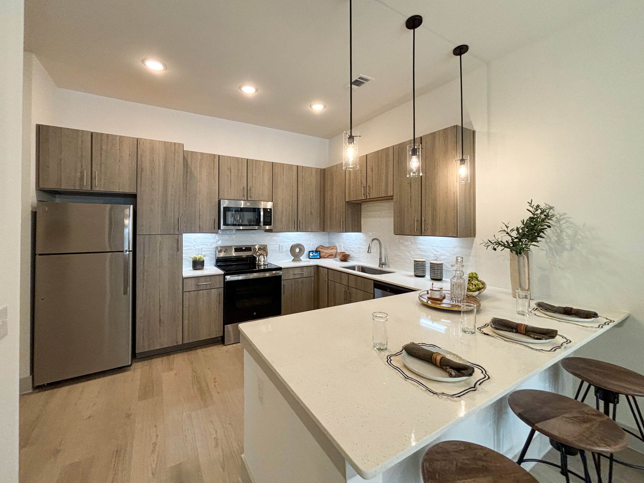 A modern northwest san antonio apartment kitchen featuring sand wooden cabinetry with a light finish, stainless steel appliances, and a white countertop. The kitchen includes a refrigerator, microwave, and an oven. The flooring is light wood, enhancing the contemporary aesthetic.