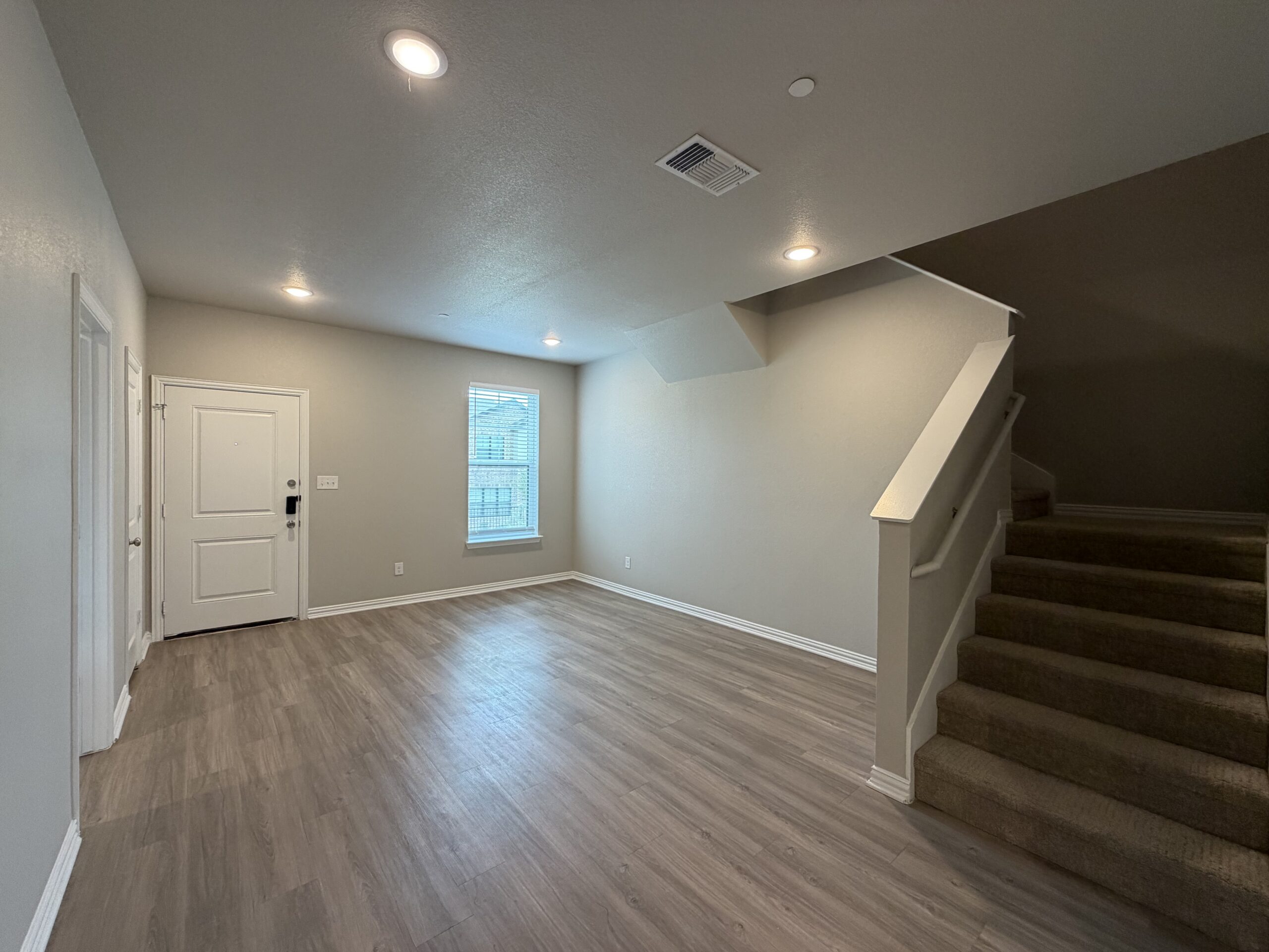 A spacious, well-lit living area features light-colored walls and a wood-like floor. To the left, there is a white front door with a security lock. A window with blinds allows natural light to enter, illuminating the room. In the background, a staircase with a white railing leads to an upper level, and the overall ambiance is modern and inviting.