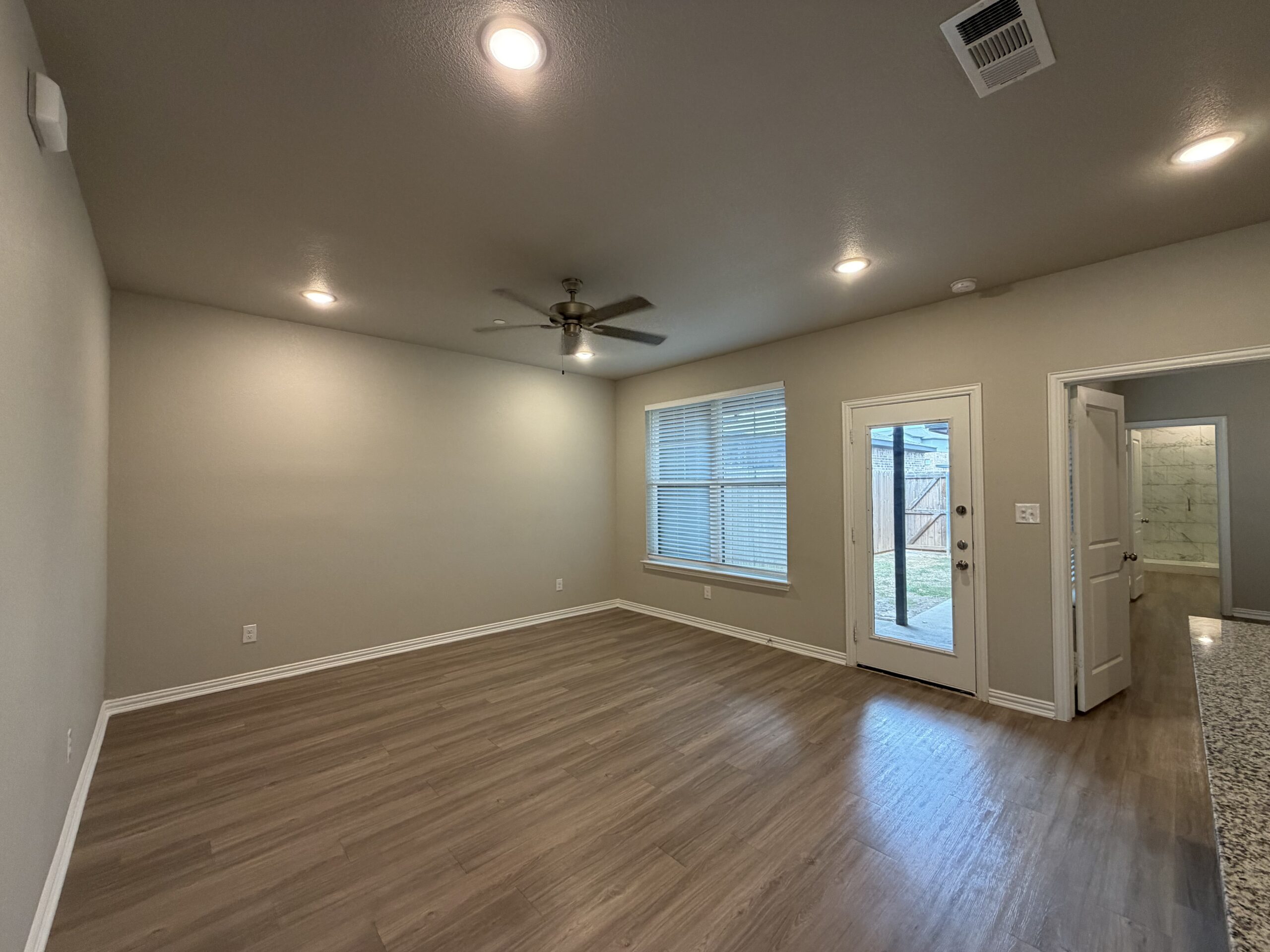 A spacious room features light-colored walls and a wood-like floor. A ceiling fan is mounted in the center, and there are recessed lights providing illumination. A large window with blinds allows natural light to enter, and a door leads outside to a patio area. Another door on the right opens to a bathroom with tiled walls visible in the background. The overall ambiance is bright and inviting.