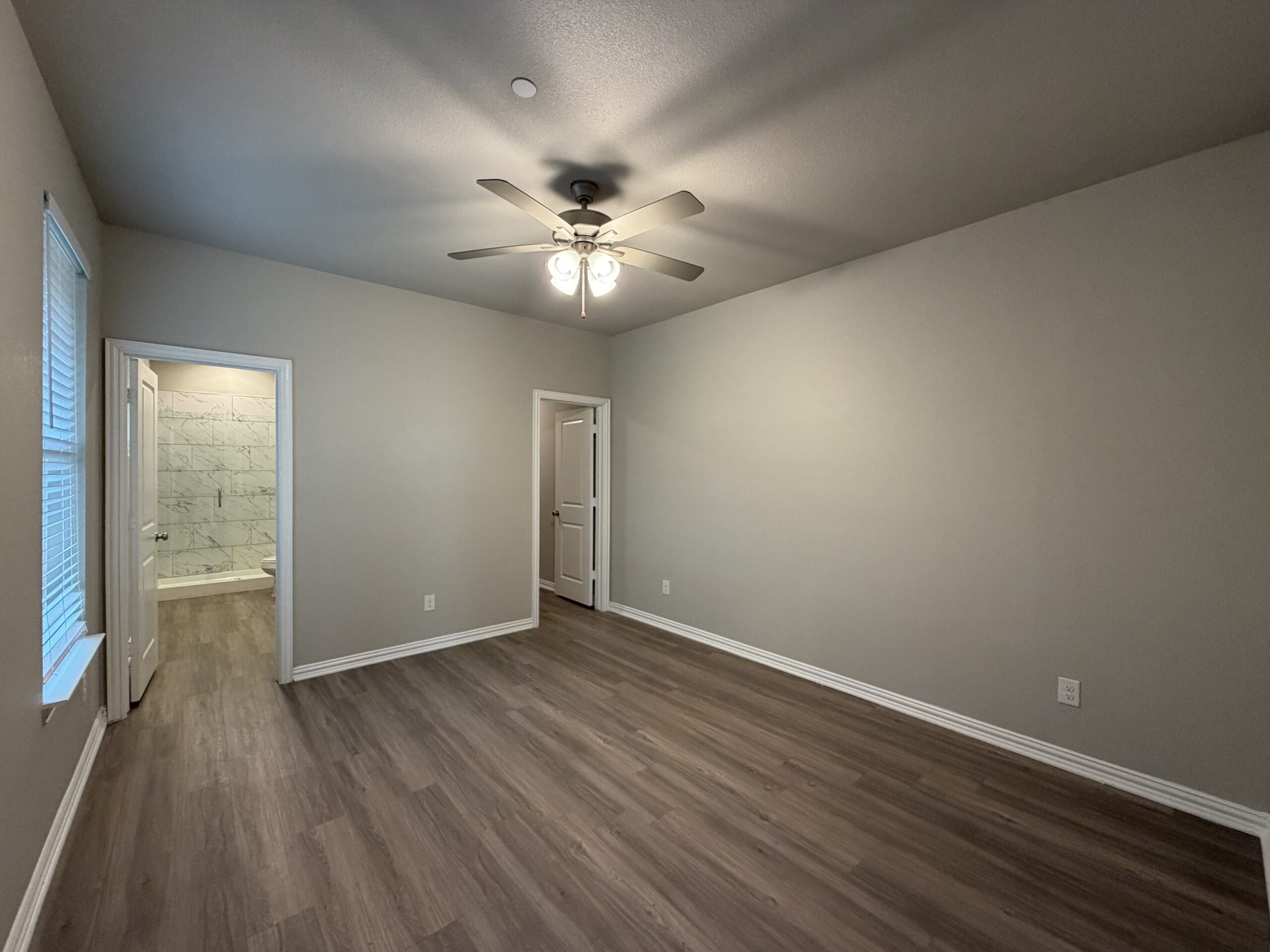 A spacious room with light gray walls and a ceiling fan featuring five bulbs. The floor is covered with light wood laminate. On the left, there is a window with blinds allowing natural light to enter. To the right, a doorway leads to another room, which appears to have a bathroom with marble-patterned tiles visible through the open door. The overall ambiance is bright and inviting, with a clean and modern aesthetic.