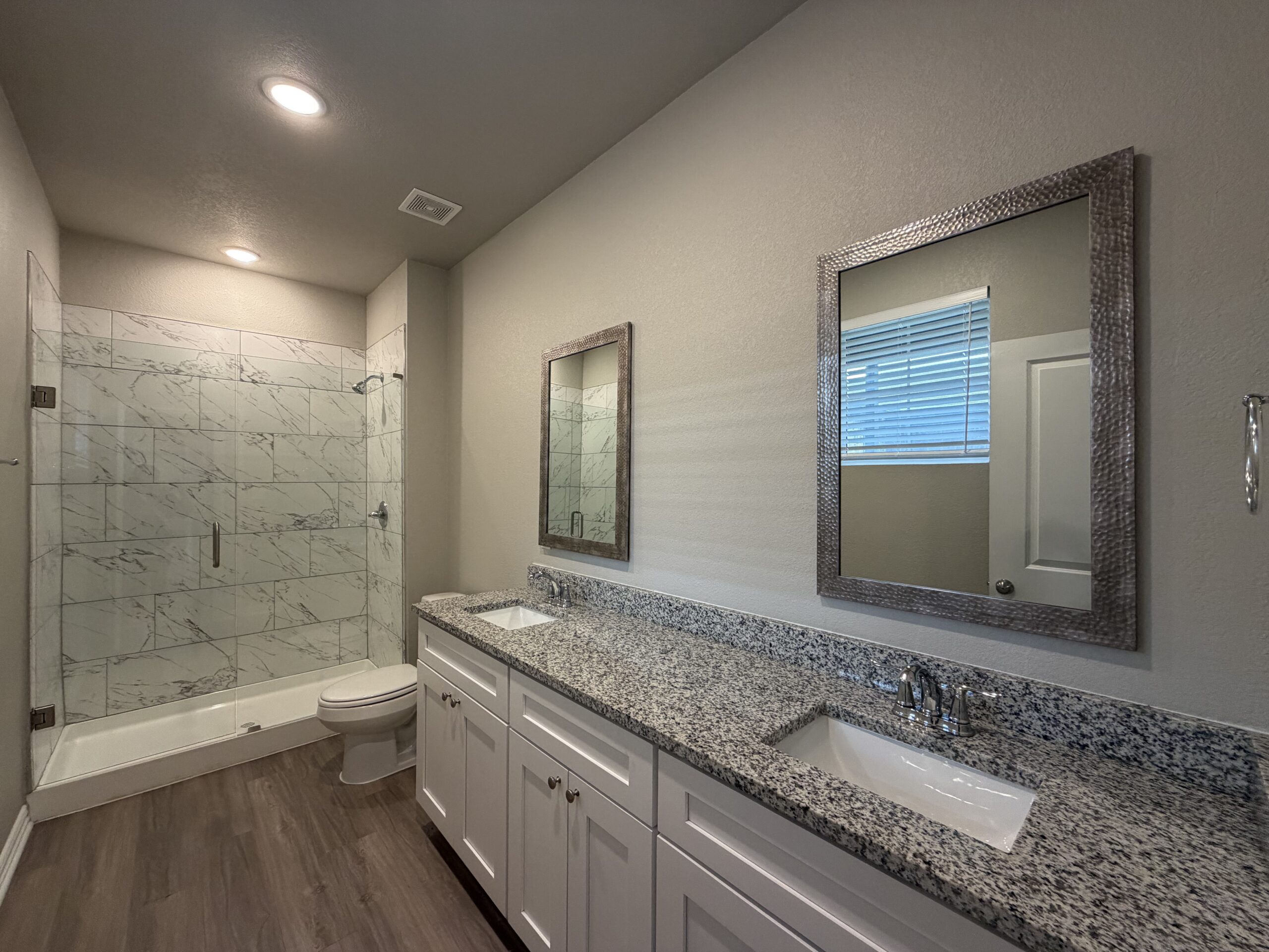 A modern bathroom features a spacious double vanity with a speckled granite countertop and two sinks. Above the vanity are two rectangular mirrors with textured frames. To the left, a glass-enclosed shower showcases marble-patterned tiles. A white toilet is positioned beside the shower, and a small window with blinds allows natural light into the space. The flooring is a light wood finish, contributing to a clean and contemporary aesthetic.